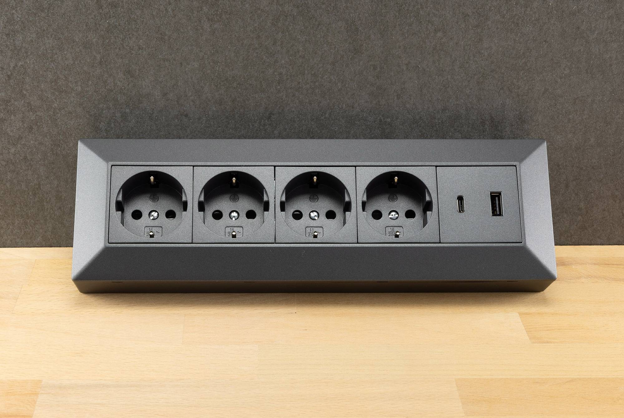 Eine Steckerleiste mit vier europäischen Steckdosen, einem USB-Anschluss und einer Taste auf einer Holzoberfläche; wird zum Anschluss mehrerer Geräte verwendet.
