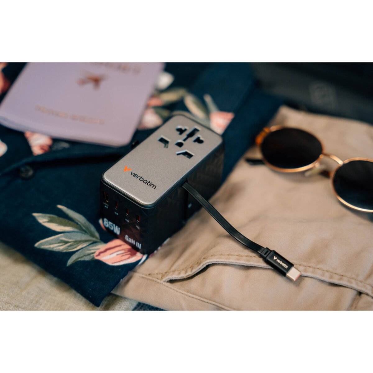 Ein Reiseadapter mit mehreren Steckdosen liegt auf einem Blumenstoff neben einer Sonnenbrille und einem geschlossenen Reisepass.