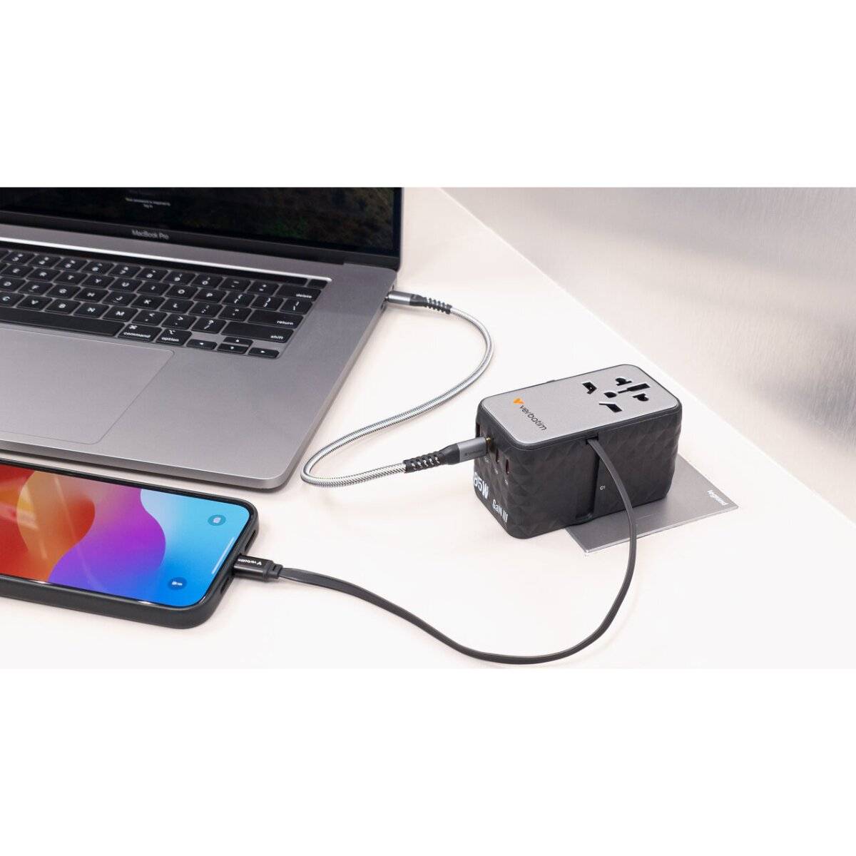 Ein Laptop und ein Smartphone sind mit einem schwarzen Reise-Stromadapter mit mehreren Steckdosenoptionen verbunden und laden sich auf einem Schreibtisch auf.