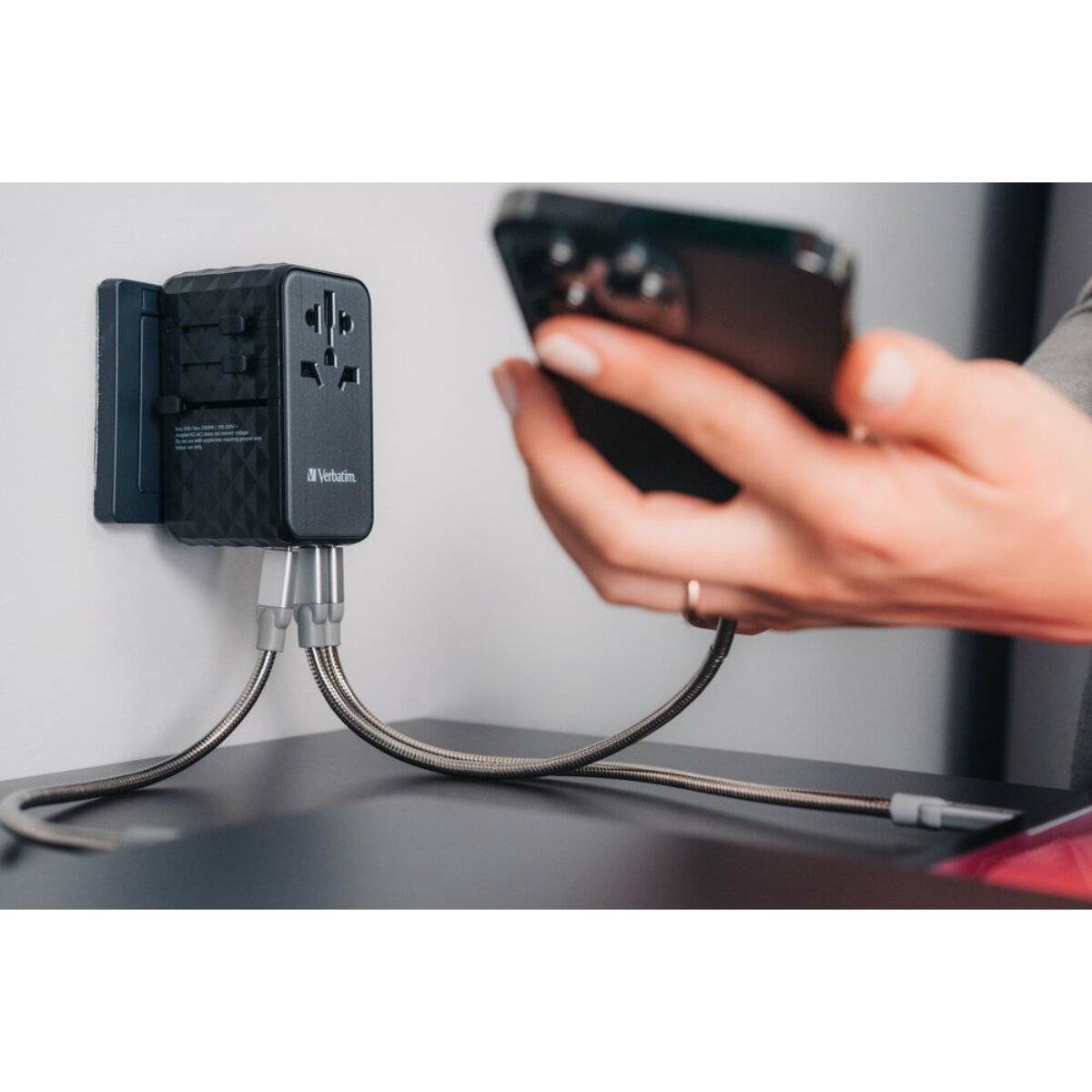 Eine Person hält ein Smartphone in der Nähe eines Verbatim-Stromadapters, der in eine Wandsteckdose eingestöpselt ist, in der Nähe von zwei Ladekabeln auf einem Tisch.