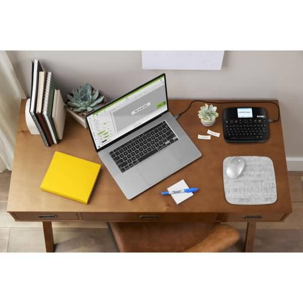 Draufsicht auf einen ordentlichen Schreibtisch eines Homeoffice mit geöffnetem Laptop, Notizbüchern, Pflanzen, einer Maus und einem Etikettendrucker auf einem Holztisch.