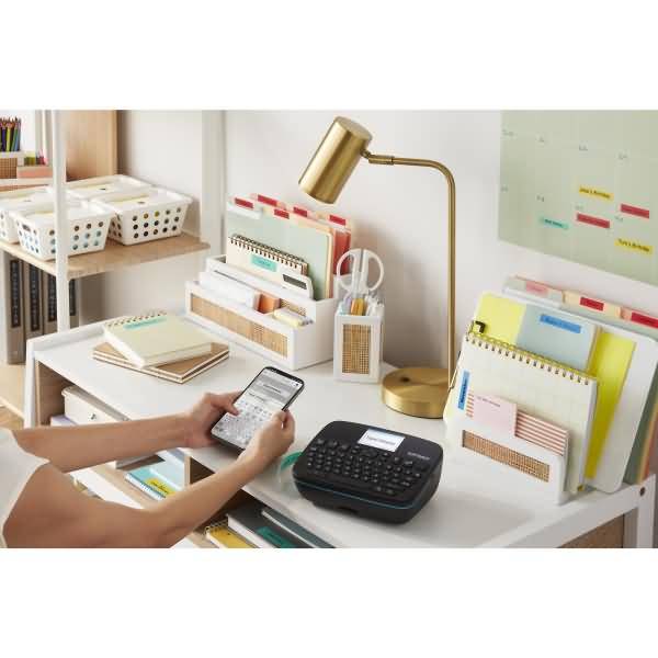 Person nutzt ein Smartphone an einem aufgeräumten Schreibtisch mit einer Etikettenmaschine, einer Lampe, Organizer und Ordnern in einem Heimbüro-Arbeitsbereich.