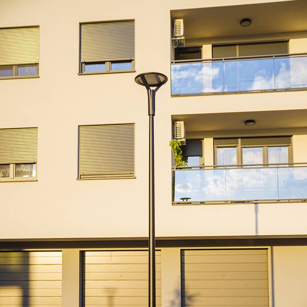 Apartmentgebäude mit geschlossenen Rollläden, Glasbalkonen und einer hohen Straßenleuchte, die Schatten im Sonnenlicht wirft.