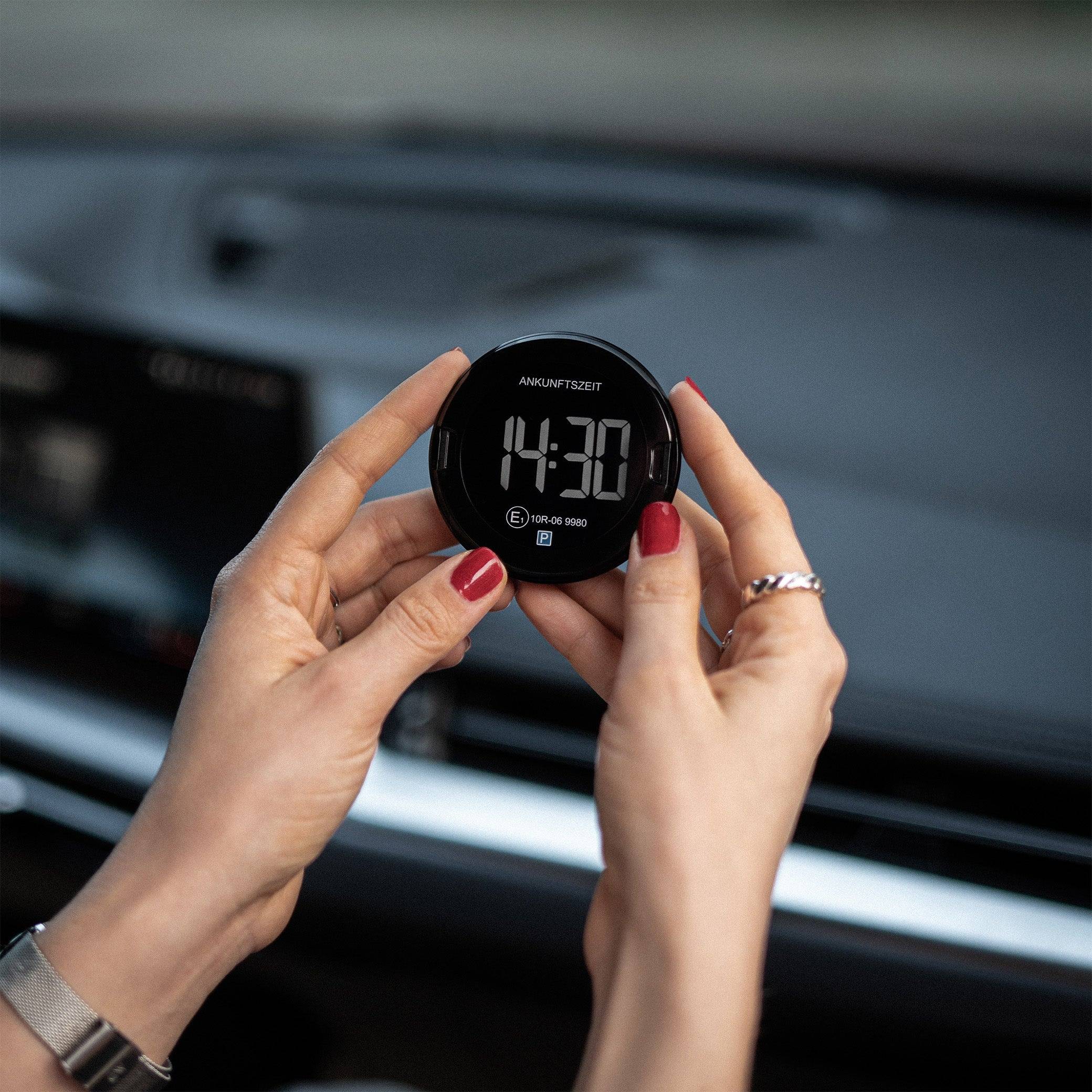 Hände halten eine kleine schwarze digitale Parkuhr, die 14:30 anzeigt, im Armaturenbrett eines Autos, verwendet um die Ankunftszeit anzuzeigen.