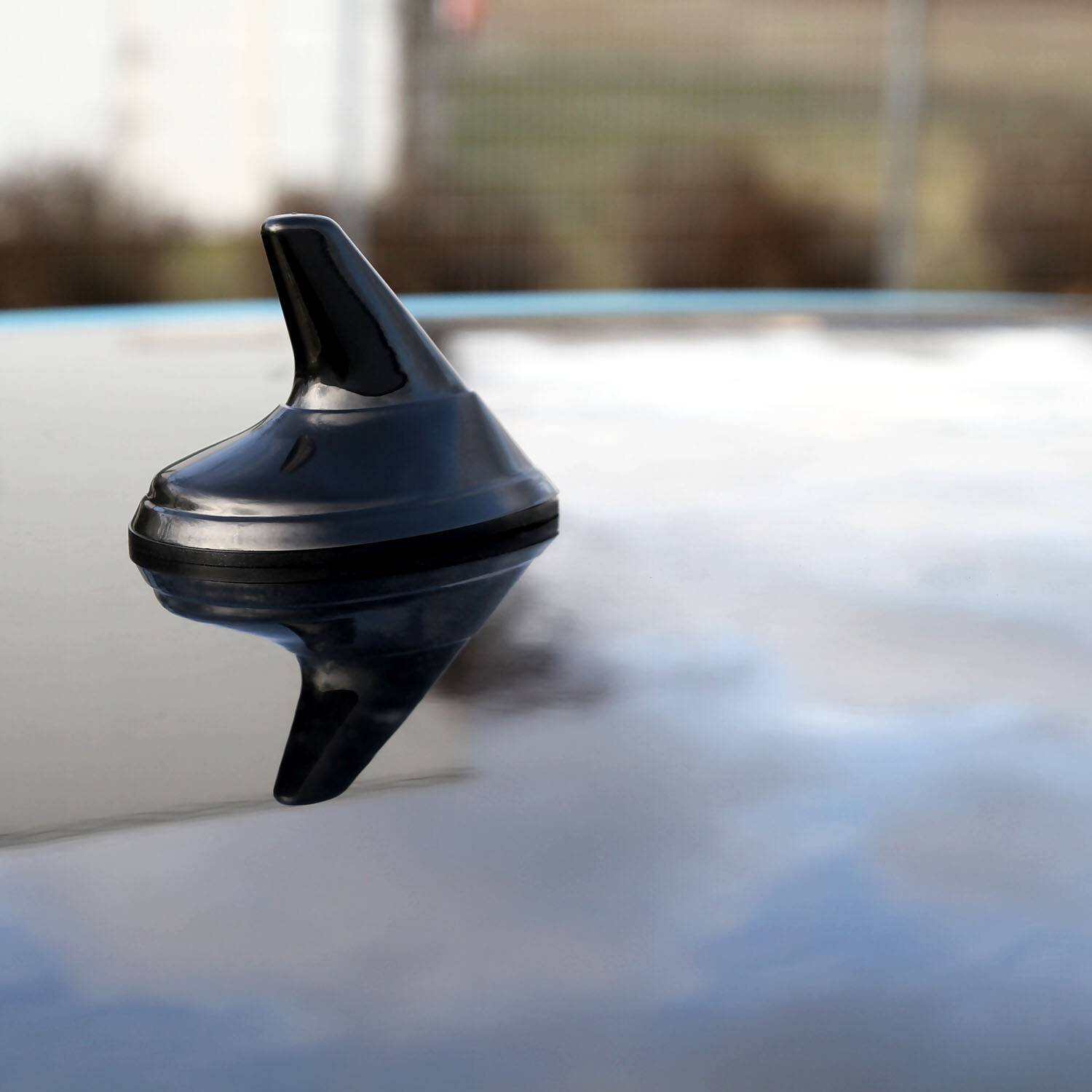 Eine kleine, schwarze, flossenfömige Antenne an einem Auto, auf einem Autodach montiert, die den bewölkten Himmel darüber widerspiegelt.