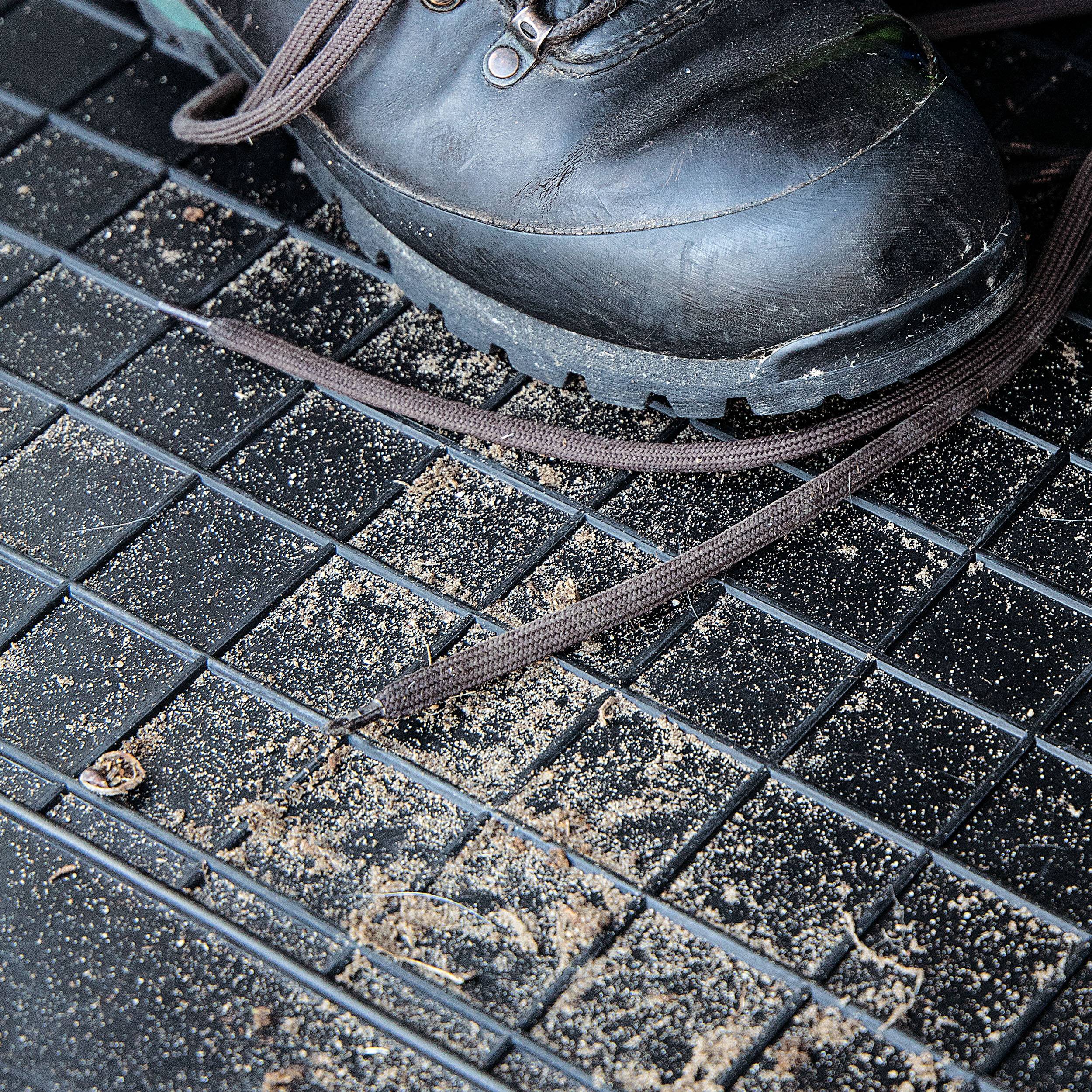 Ein schmutziger schwarzer Stiefel mit braunen Schnürsenkeln auf einer strukturierten schwarzen Matte, bedeckt mit Schmutz, was auf Outdoor-Aktivitäten oder Wandern hindeutet.