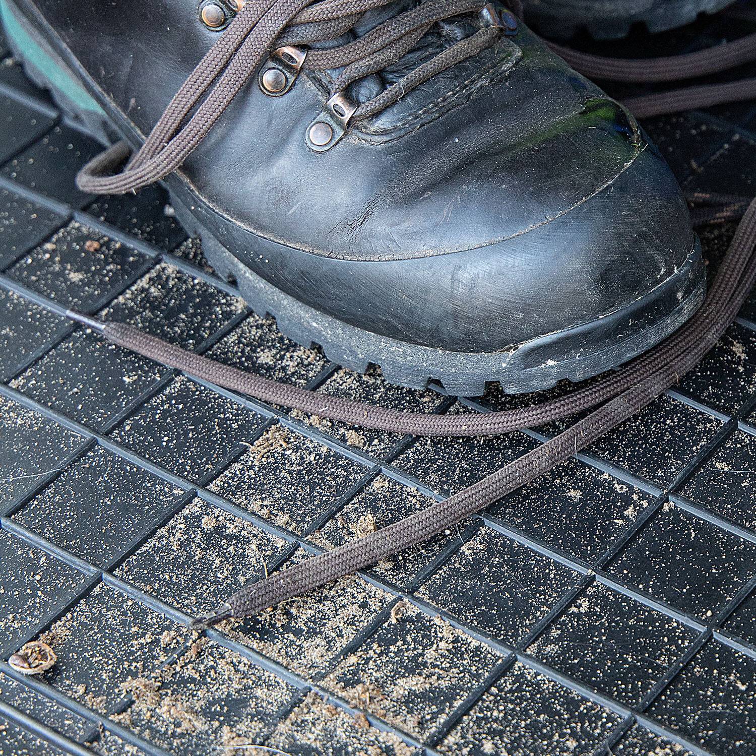 Nahaufnahme eines schwarzen Stiefels mit langen Schnürsenkeln auf einer schmutzigen, gittergemusterten Bodenmatte, die auf Outdoor- oder raue Nutzung hindeutet.