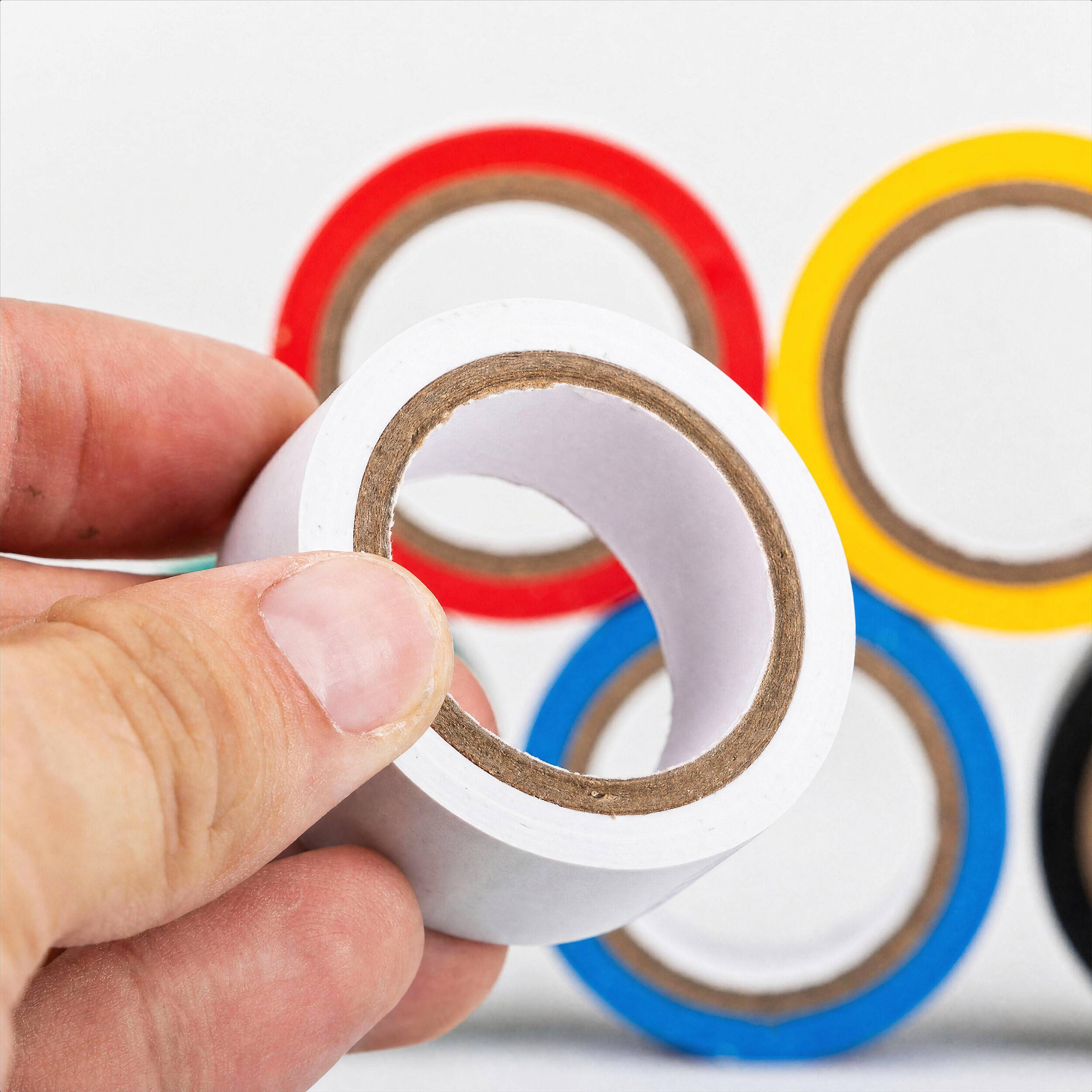 Hand hält eine Rolle weißes Klebeband vor roten, gelben und blauen Klebeband-Rollen auf weißem Hintergrund.