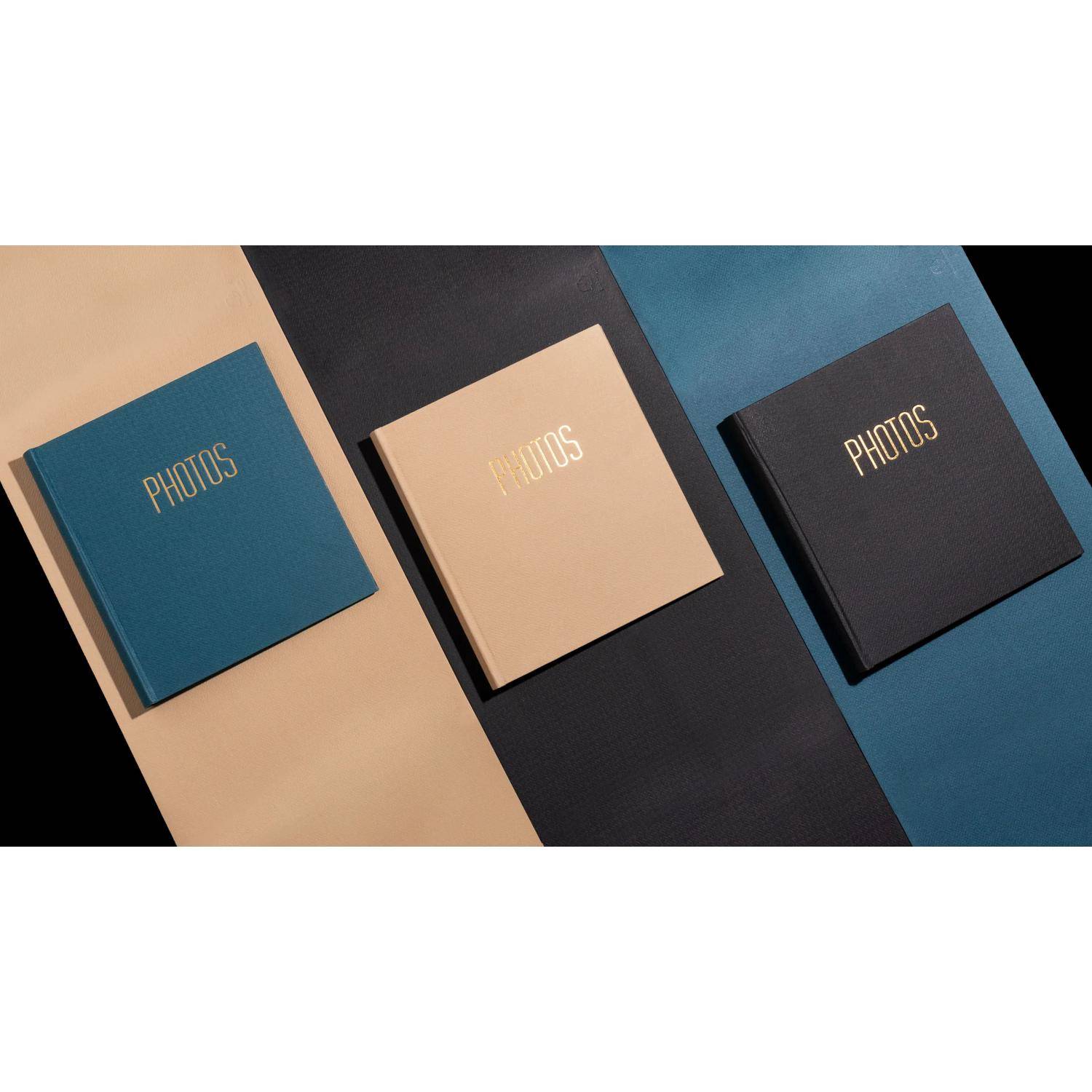 Drei quadratische Fotoalben mit der Aufschrift „PHOTOS" in Petrol, Beige und Schwarz, angeordnet auf geneigten blauen, beige- und schwarzfarbenen Paneelen.
