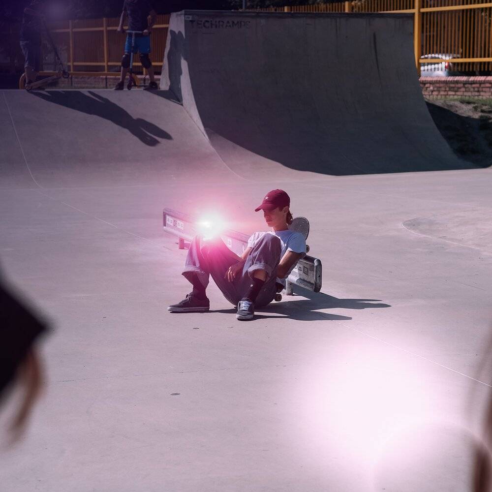 Eine Person sitzt auf dem Boden in einem Skatepark und hält eine Lichtquelle. Ein Skateboard ist in der Nähe und weitere befinden sich im Hintergrund.