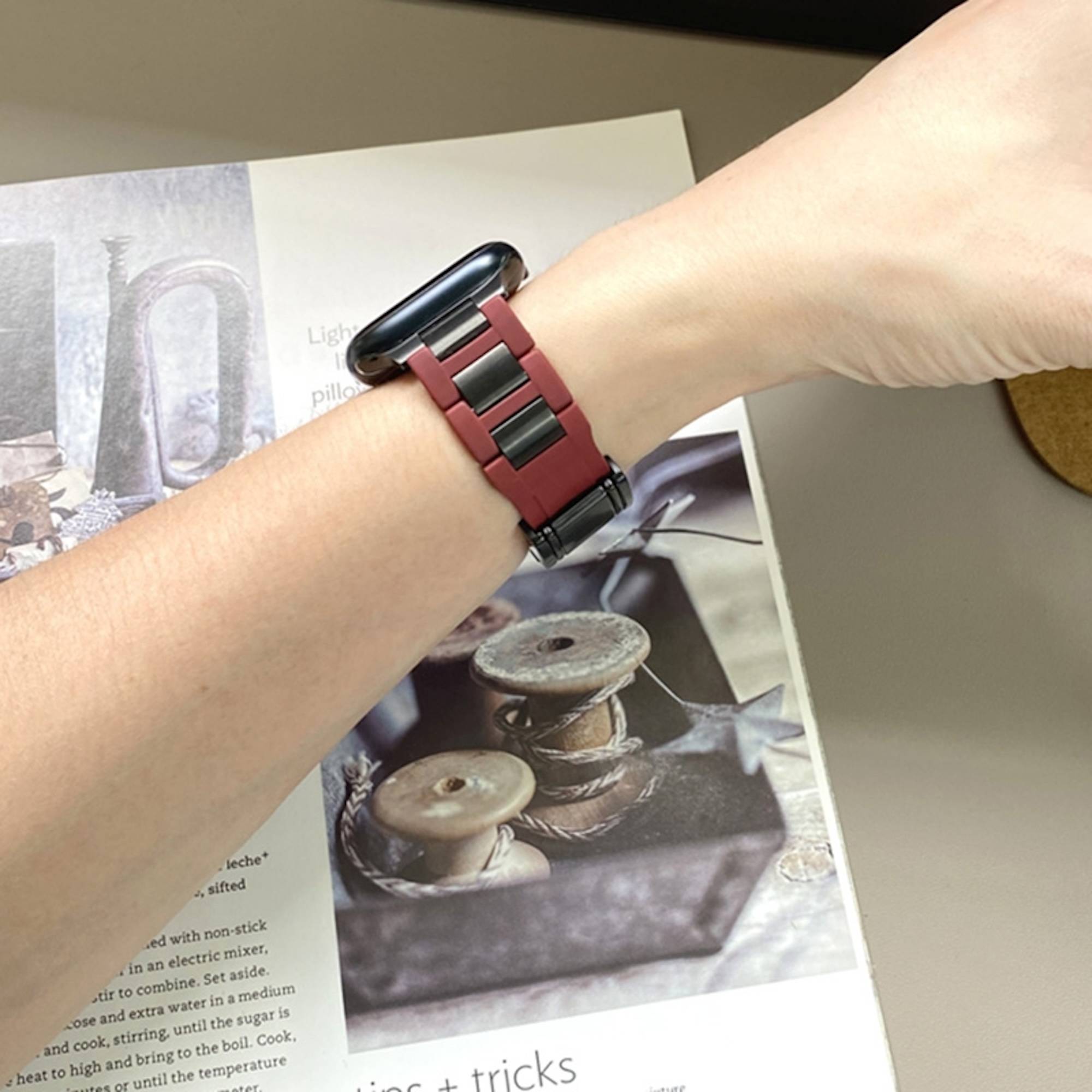 Person mit einer rot-schwarzen Smartwatch mit schwarzem rechteckigem Bildschirm, gezeigt auf einem ausgestreckten Handgelenk über einer Magazinseite.