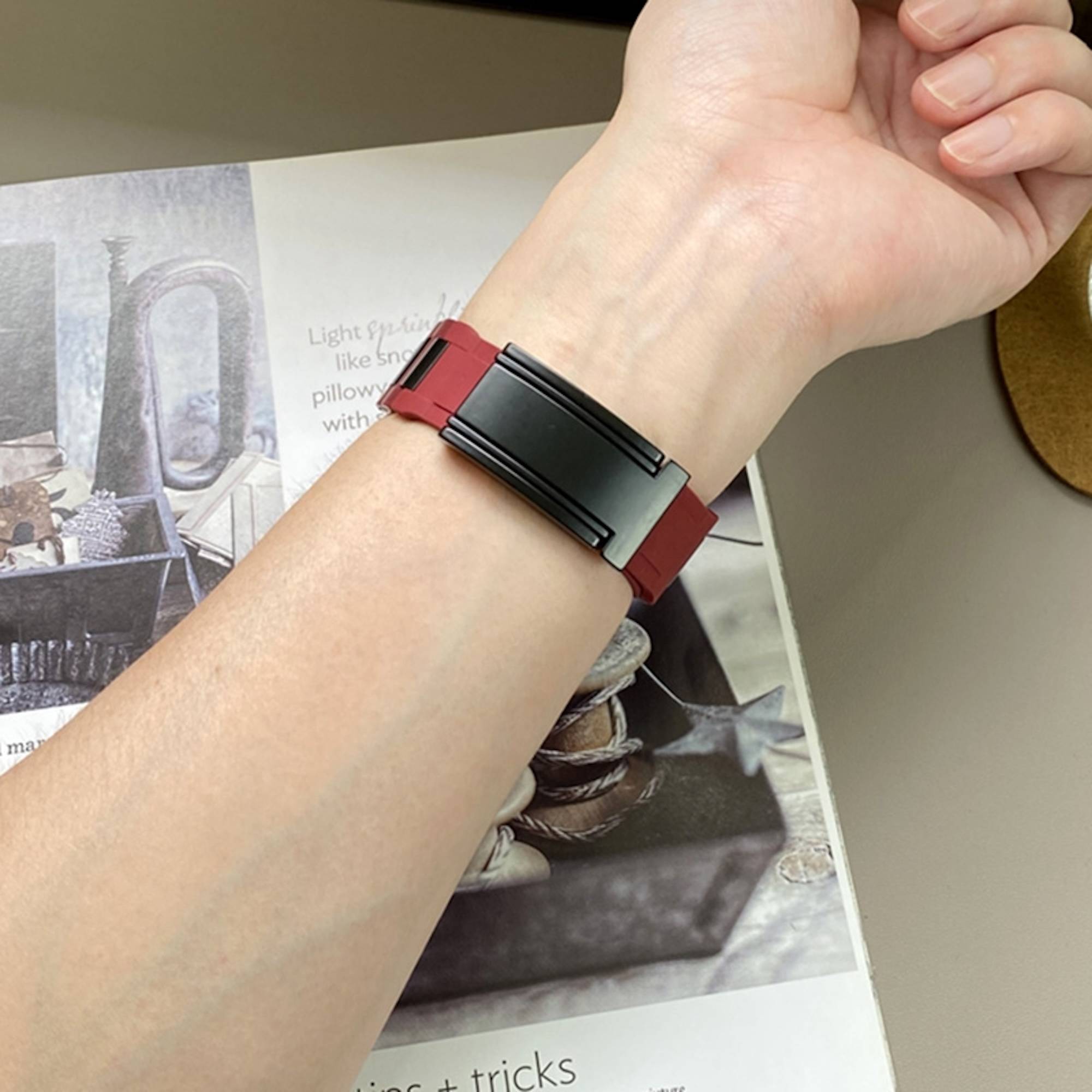 Handgelenk mit einem schwarzroten Fitness-Tracker-Band über einem offenen Magazin auf einem Tisch.
