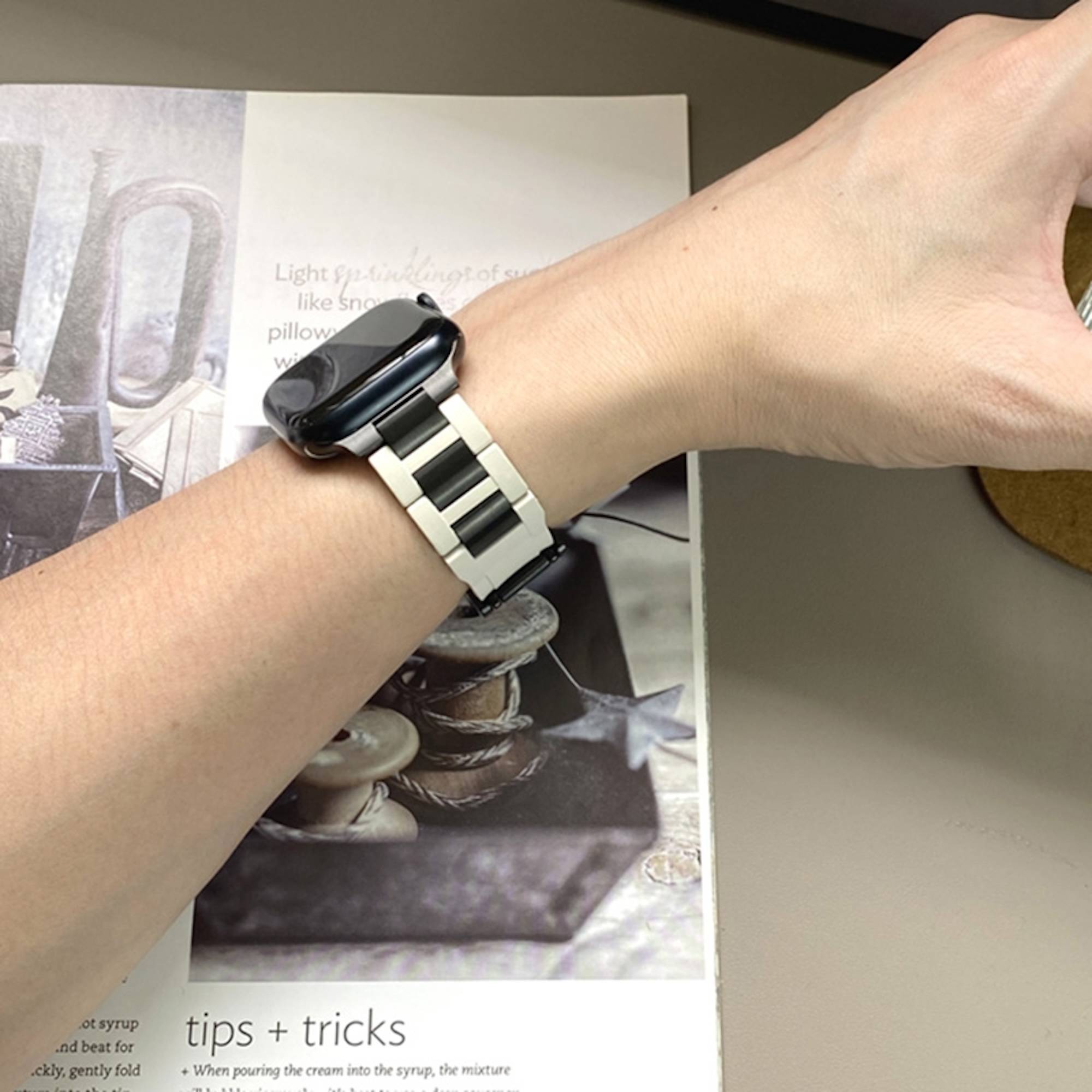 Eine Person, die eine schwarze Smartwatch mit einem silbernen Metallarmband am Handgelenk über einer aufgeschlagenen Magazinseite trägt.