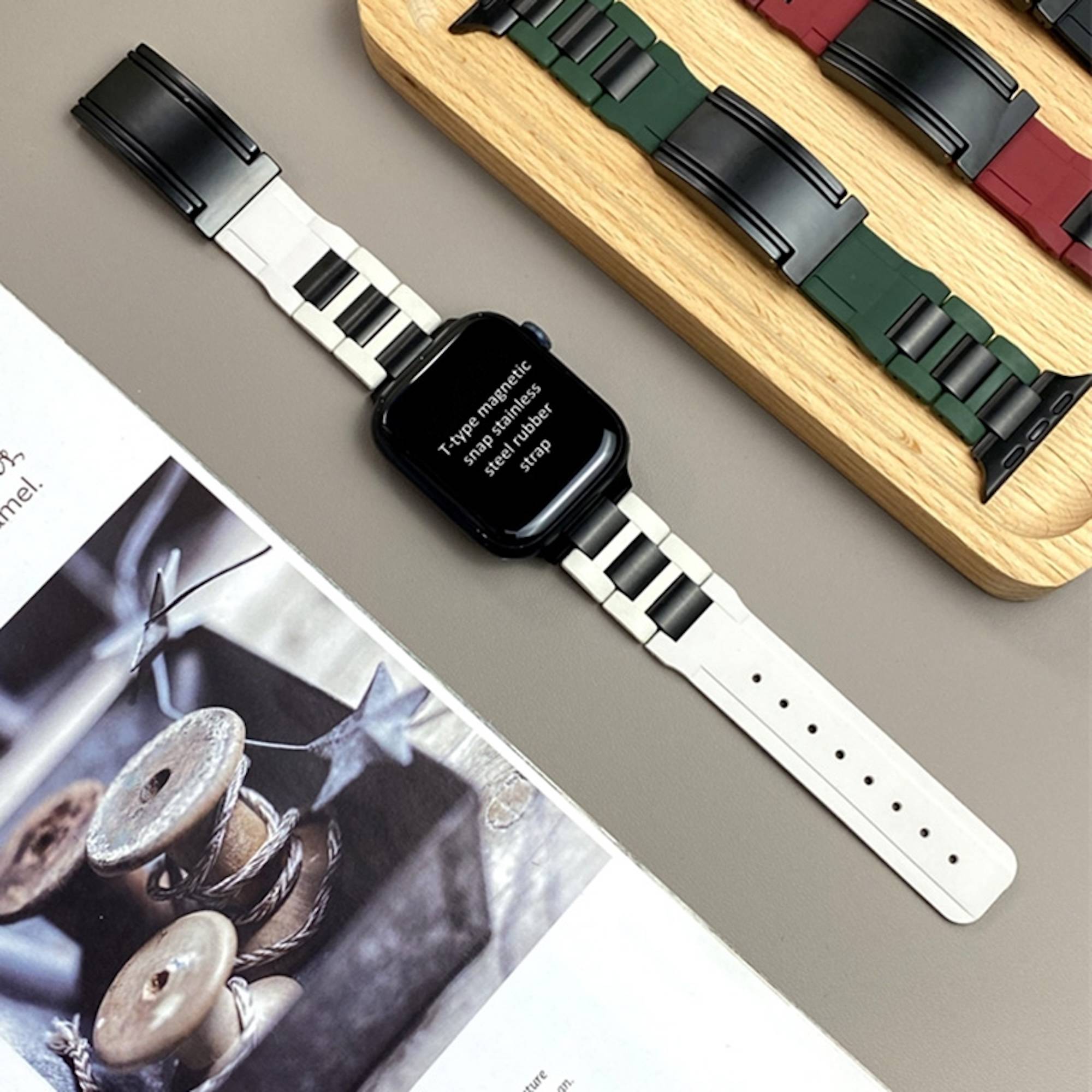 Smartwatch mit schwarzem quadratischem Zifferblatt und weiß-schwarzem Magnetarmband, gezeigt neben zwei alternativen Uhrenarmbändern auf einem Holztablett und einer Magazinseite.