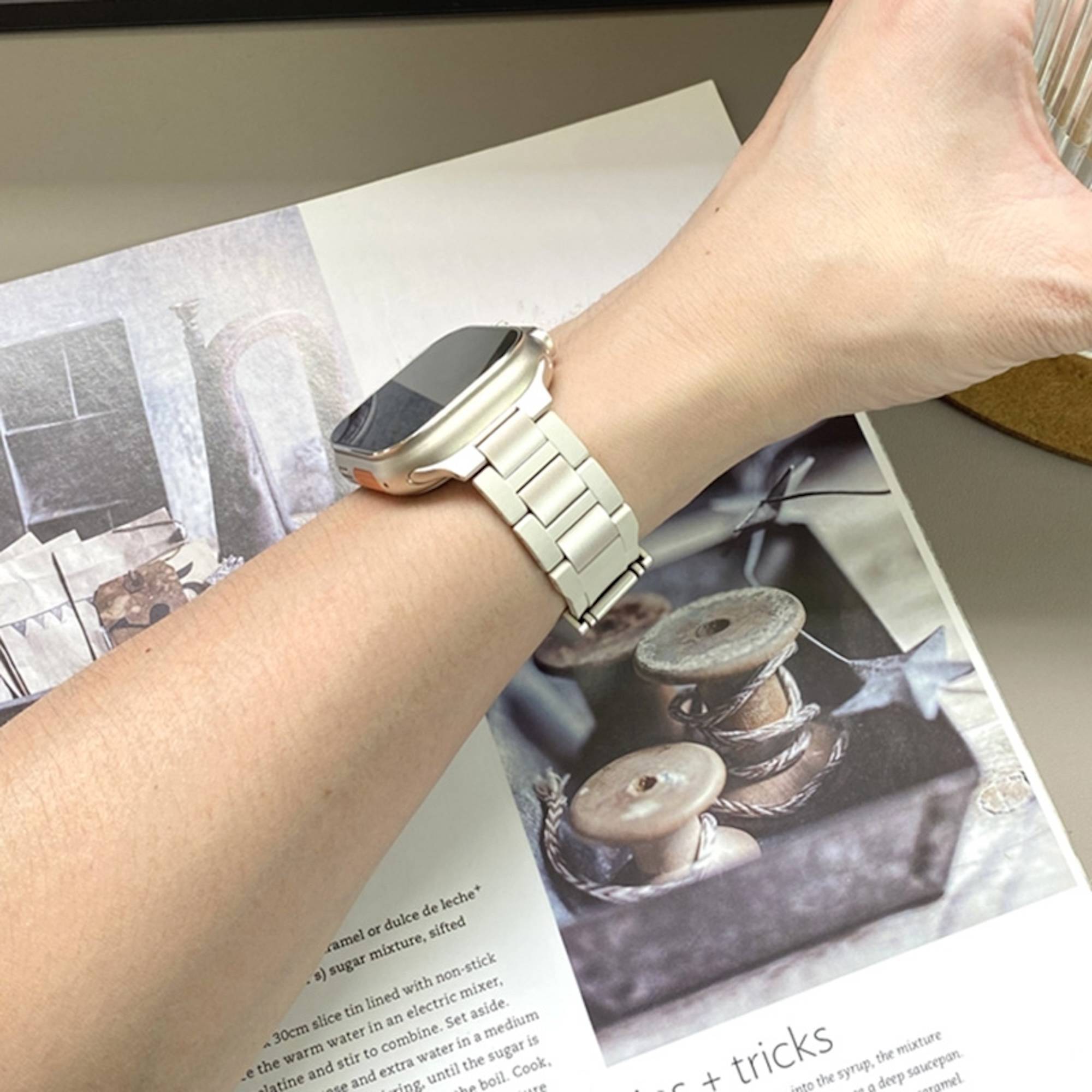 Person trägt eine silberne Smartwatch mit Metallgliedband auf einer offenen Magazinseite mit Handwerk-Spulen-Bildmotiven.