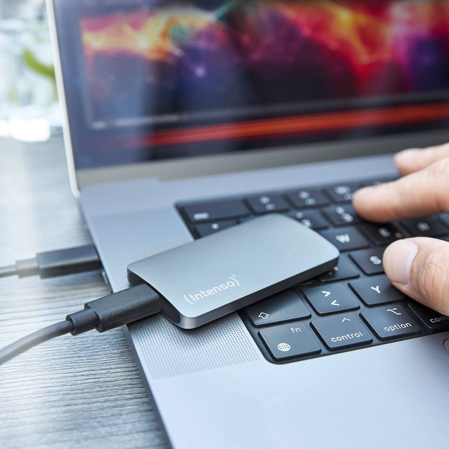 Eine Hand schließt eine externe Festplatte an einen Laptop an. Der Bildschirm zeigt bunte Grafiken. Die Situation deutet auf Datenübertragung oder Datensicherung hin.