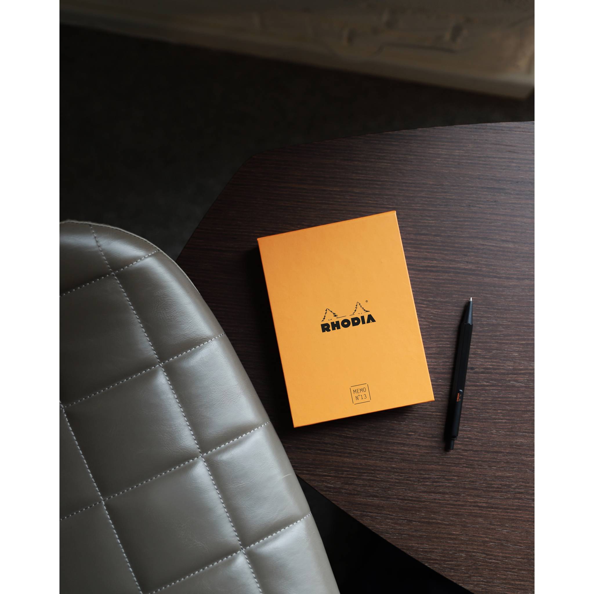 Oranges Rhodia-Notizbuch und schwarzer Stift auf einem dunklen Holzschreibtisch neben einem grauen Lederstuhl.