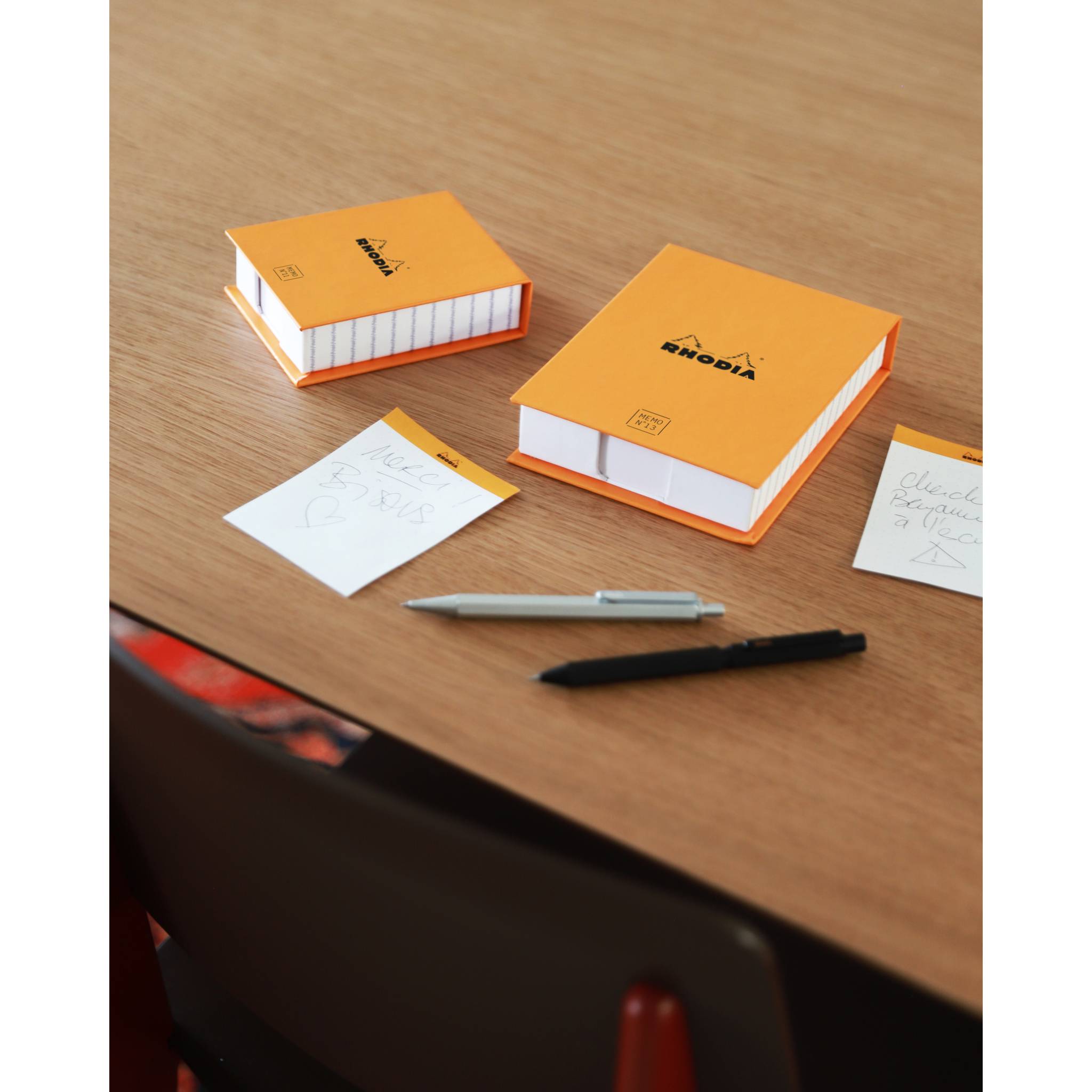 Zwei orangefarbene Rhodia-Notizblöcke auf einem Holzschreibtisch, mit Stiften und kleinen Notizkarten in der Nähe.