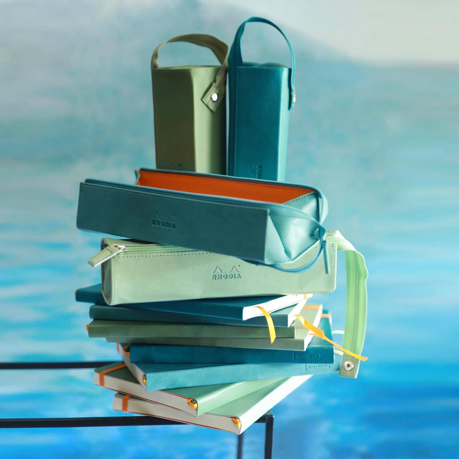 Stapel farbenfroher Rhodia-Notizbücher und Reißverschlussetuis, wie ein Turm auf blauem Hintergrund angeordnet.
