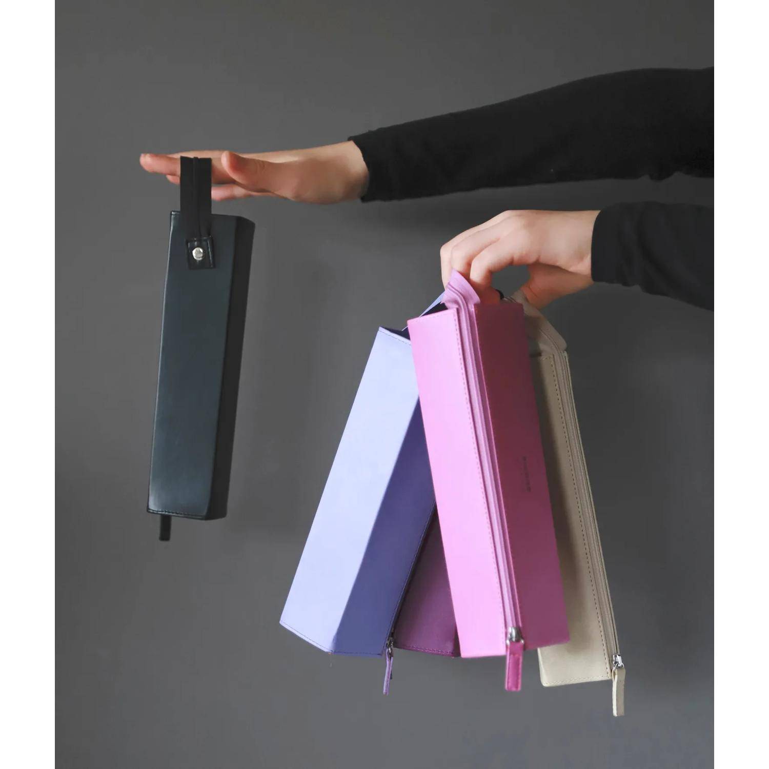 Hände halten mehrere pastellfarbene Reißverschlussbeutel oder kleine Taschen sowie ein schwarzes Etui vor einem grauen Hintergrund.