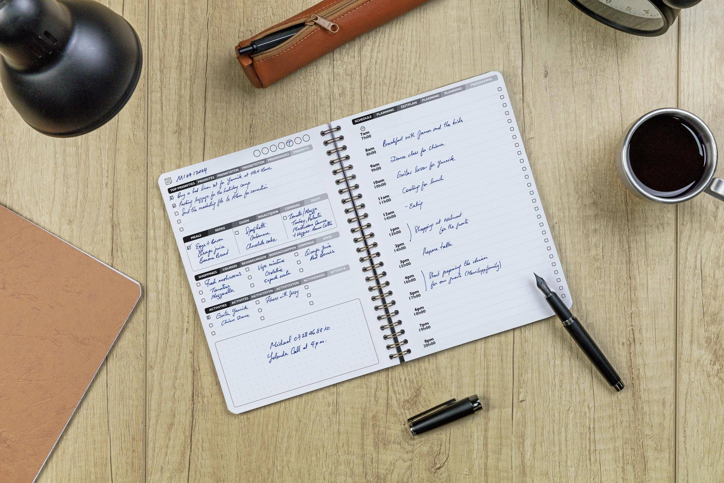 Ein offener Spiralplaner mit handschriftlichen Notizen und To-Do-Listen liegt auf einem Holzschreibtisch neben einem Stift, einer Kaffeetasse, einer Lampe und einem Notizbuch.