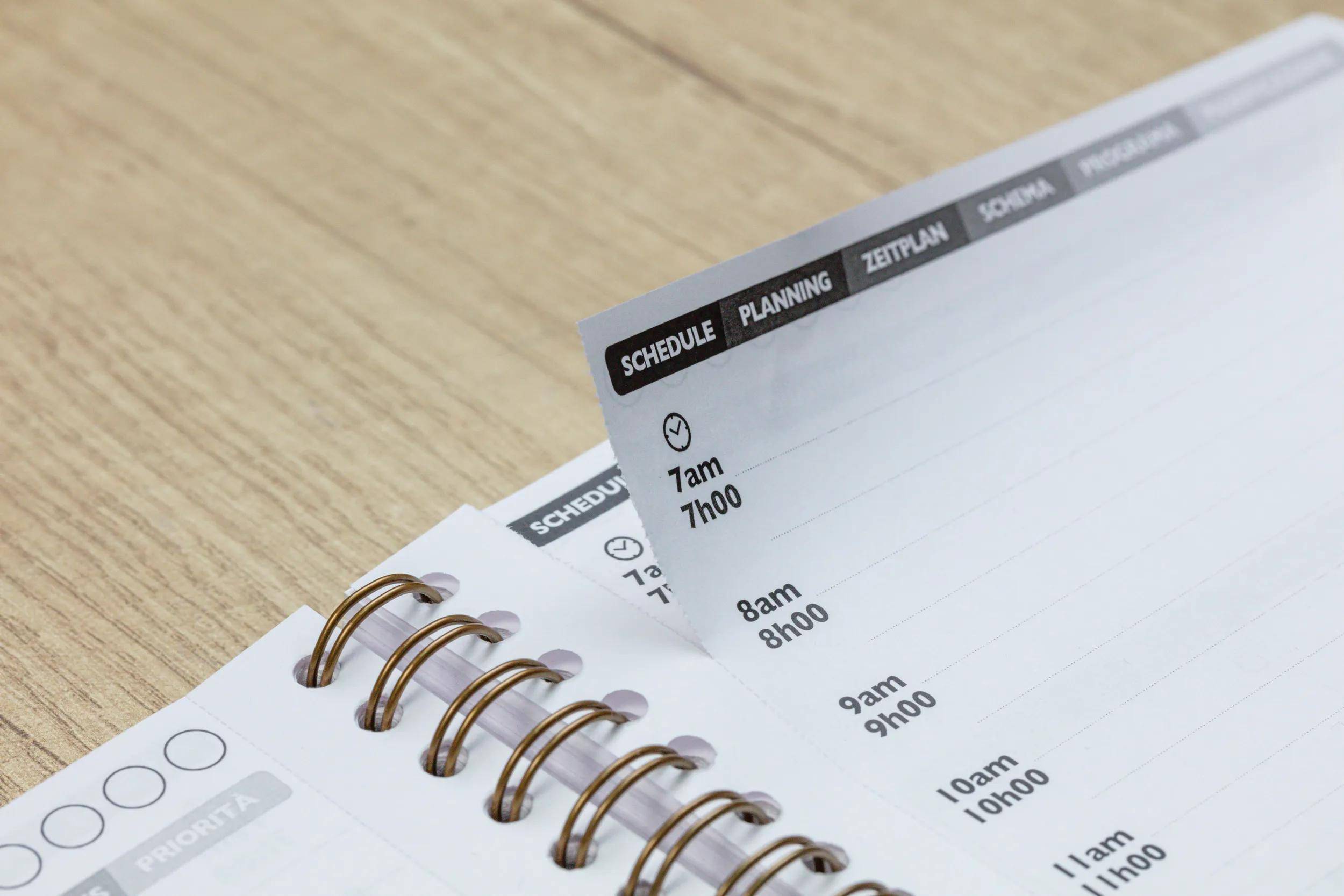 Nahaufnahme eines Plannernotizbuches mit einer Wochenschedulseite, die Zeitfenster von 7 Uhr morgens bis 11 Uhr morgens zeigt und Registerkarten für Zeitplan, Planung und andere Abschnitte aufweist.