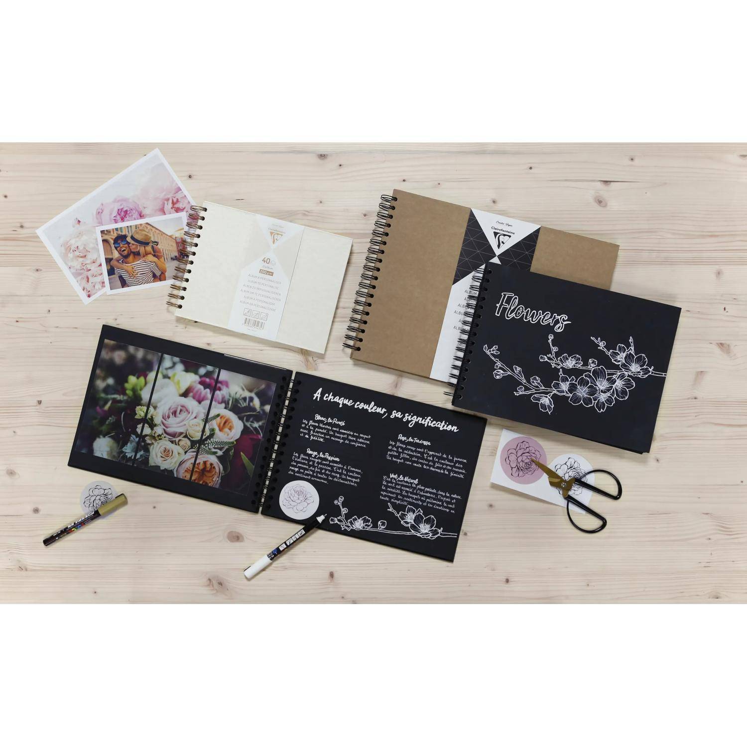 Flache Anordnung aus handgefertigten floralen Scrapbook- und Tagebuchseiten auf einem Holztisch, einschließlich schwarzer Coverdesigns, Fotoserien und Handwerkswerkzeugen.