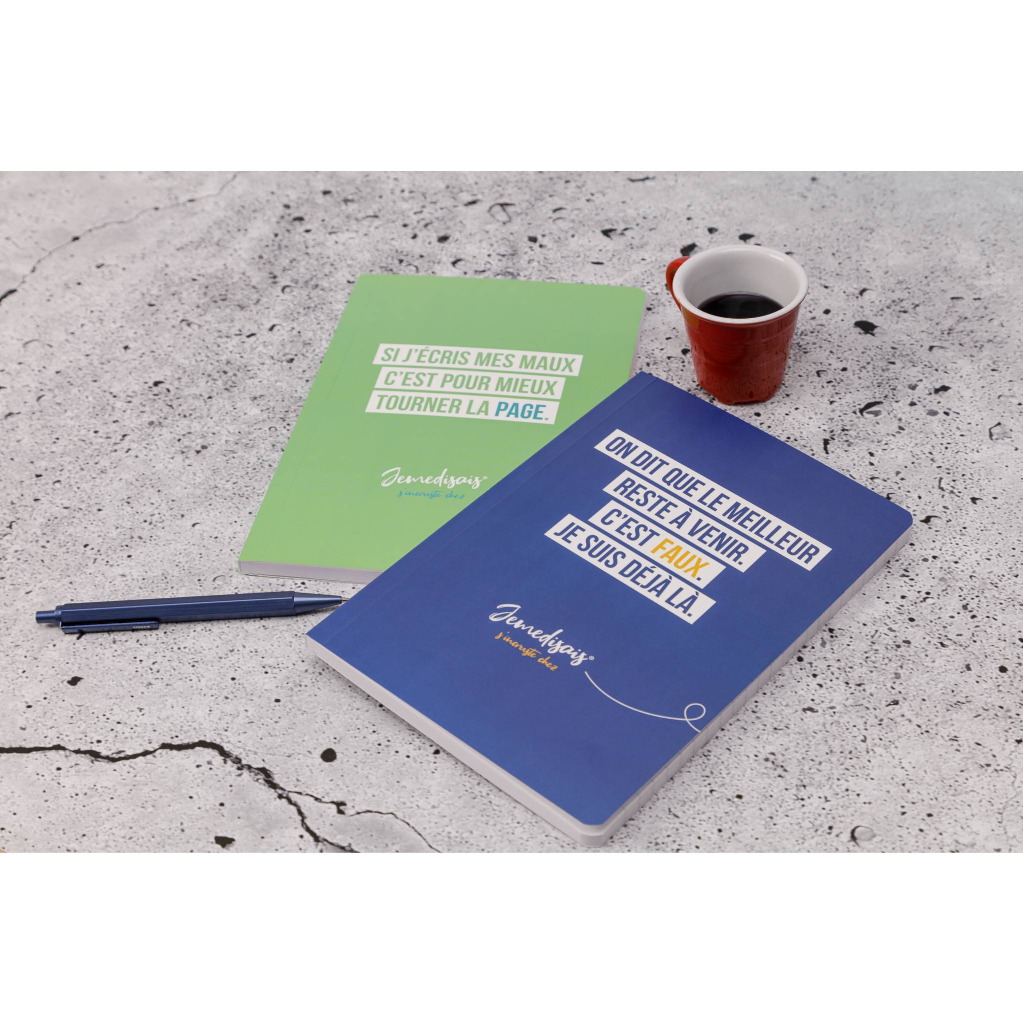 Zwei Notizbücher mit französischem Text, ein Stift und eine rote Tasse Kaffee auf einer Betonoberfläche.