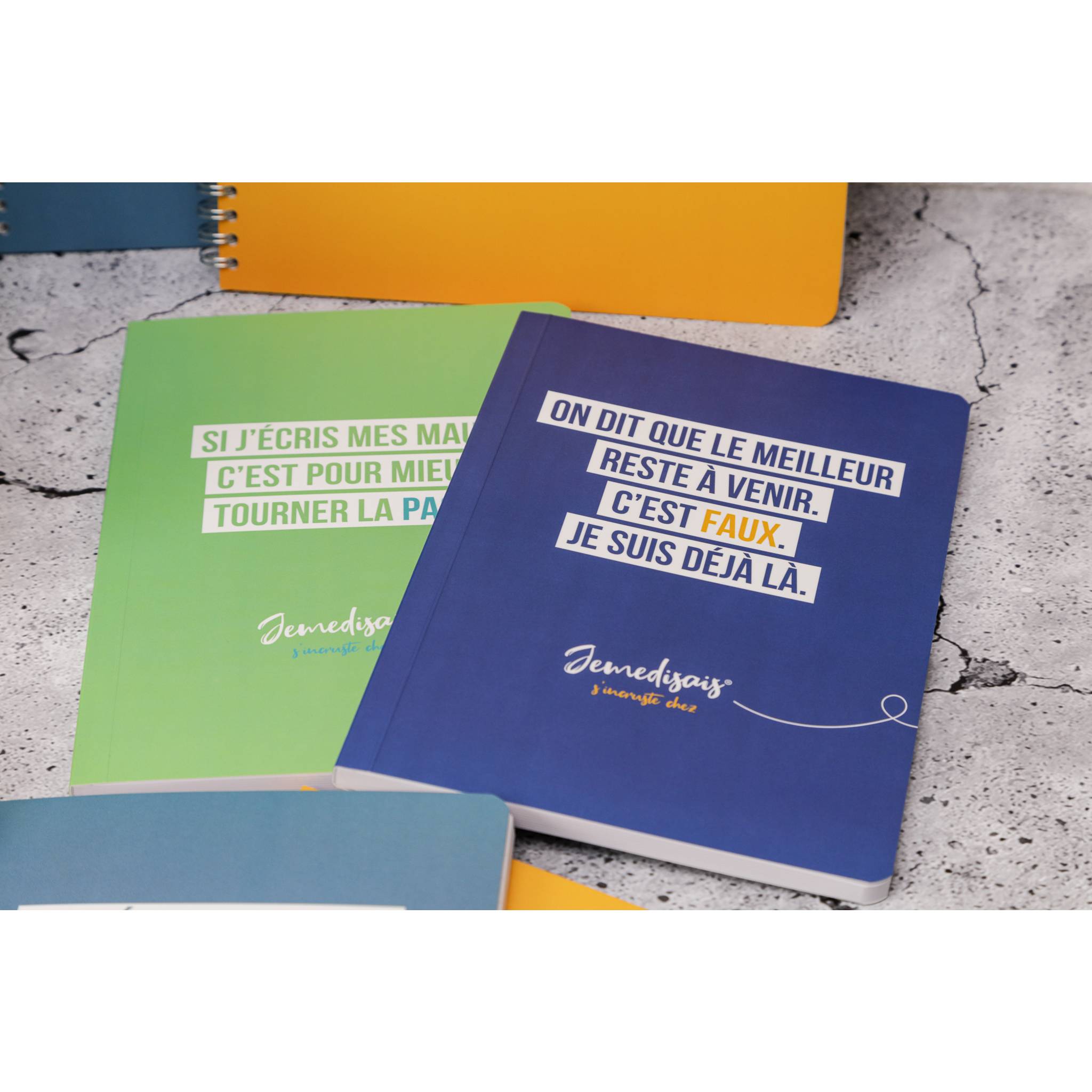 Zwei Notizbücher auf einer gesprenkelten Tischplatte: eines mit blauem Einband trägt die Aufschrift „ON DIT QUE LE MEILLEUR RESTE À VENIR. C'EST FAUX. JE SUIS DÉJÀ LÀ." und eines mit grünem Einband hat teilweise sichtbaren französischen Text.