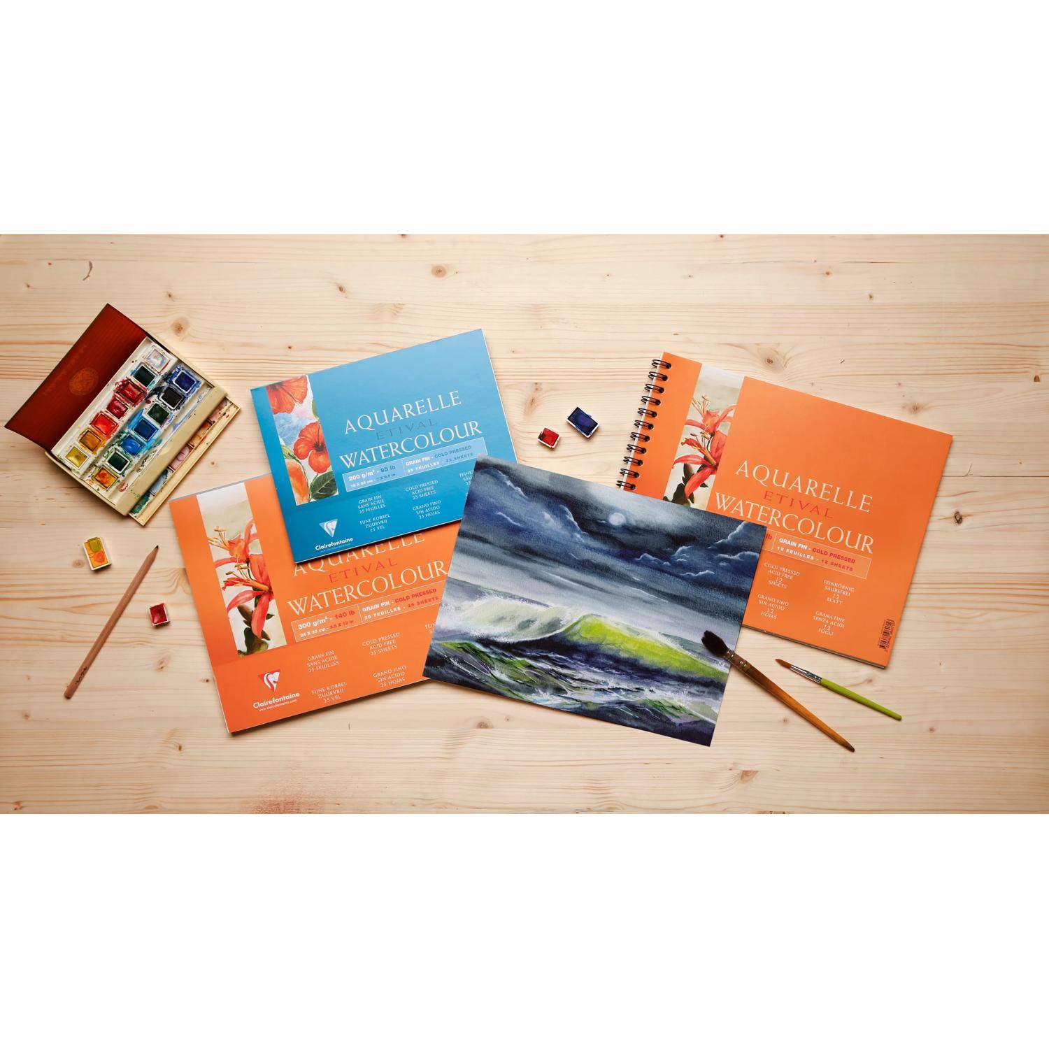 Aquarellmalerei-Materialien und Skizzenbücher auf einem Holztisch, darunter "AQUARELLE WATERCOLOUR"" Blöcke, Farben, Pinsel und ein Landschaftsgemälde.