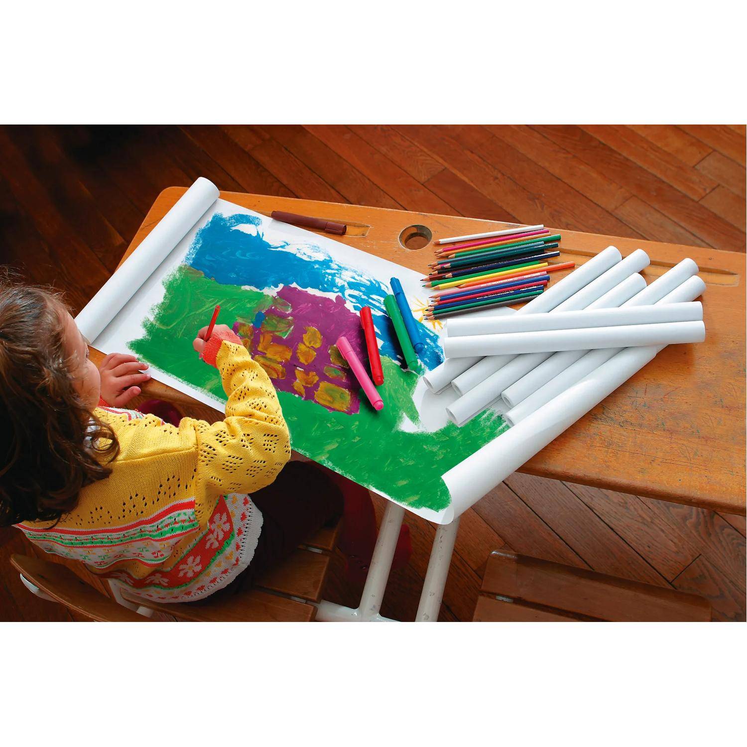 Kind zeichnet mit Buntstiften auf einer großen Papierrolle, die über einem Tisch ausgebreitet ist, mit farbigen Markern und aufgerollten Papierrollen in der Nähe.