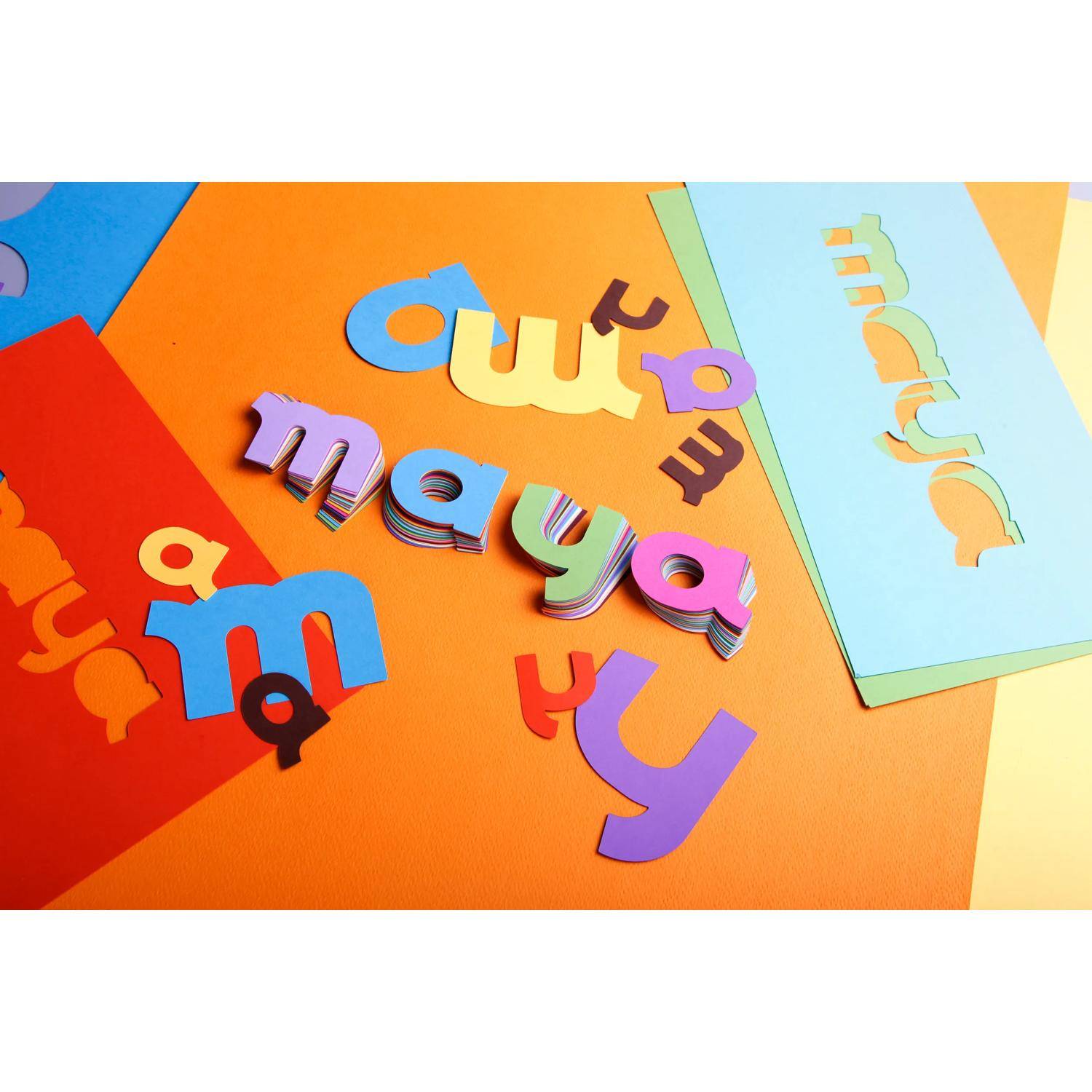 Bunte ausgeschnittene Papierbuchstaben, die „cut maya" buchstabieren, auf leuchtendem orangefarbenem Papier angeordnet, mit anderen abstrakten farbigen Blättern in der Nähe.