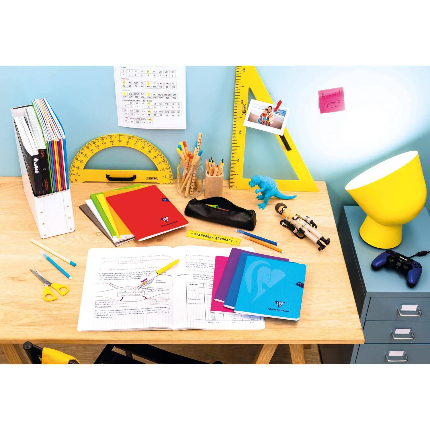 Ein unordentlicher Schreibtisch eines Studierenden mit Lehrbüchern, Notizbüchern, Stiften, Linealen, Scheren, Kalendern und kleinen Spielzeugen, neben einer Lampe und einem Spielecontroller.