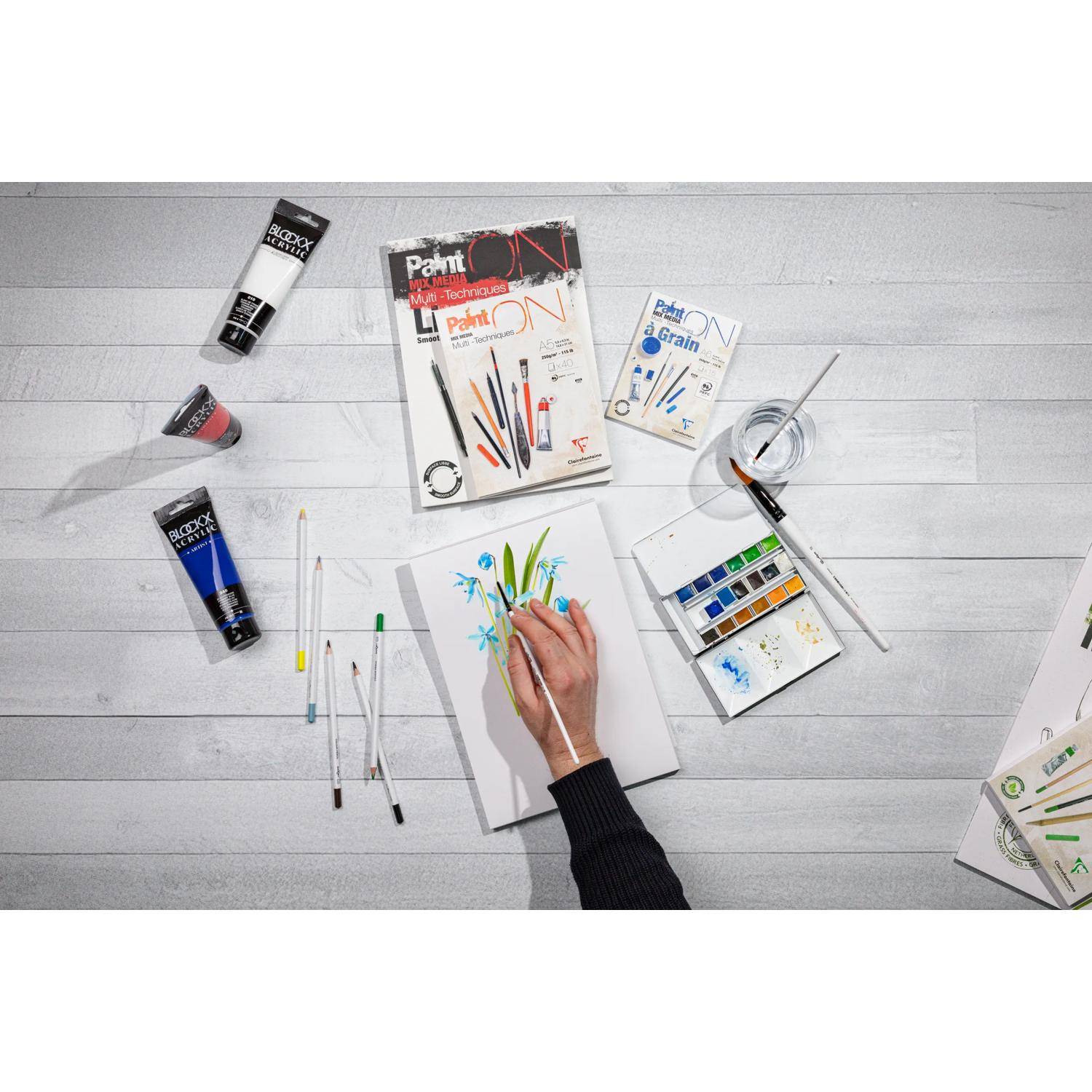 Draufsicht einer Person, die ein florales Aquarell auf Papier malt, umgeben von Farbtuben, Pinseln, Farbpaletten und Kunstmagazinen auf einem weißen Schreibtisch.