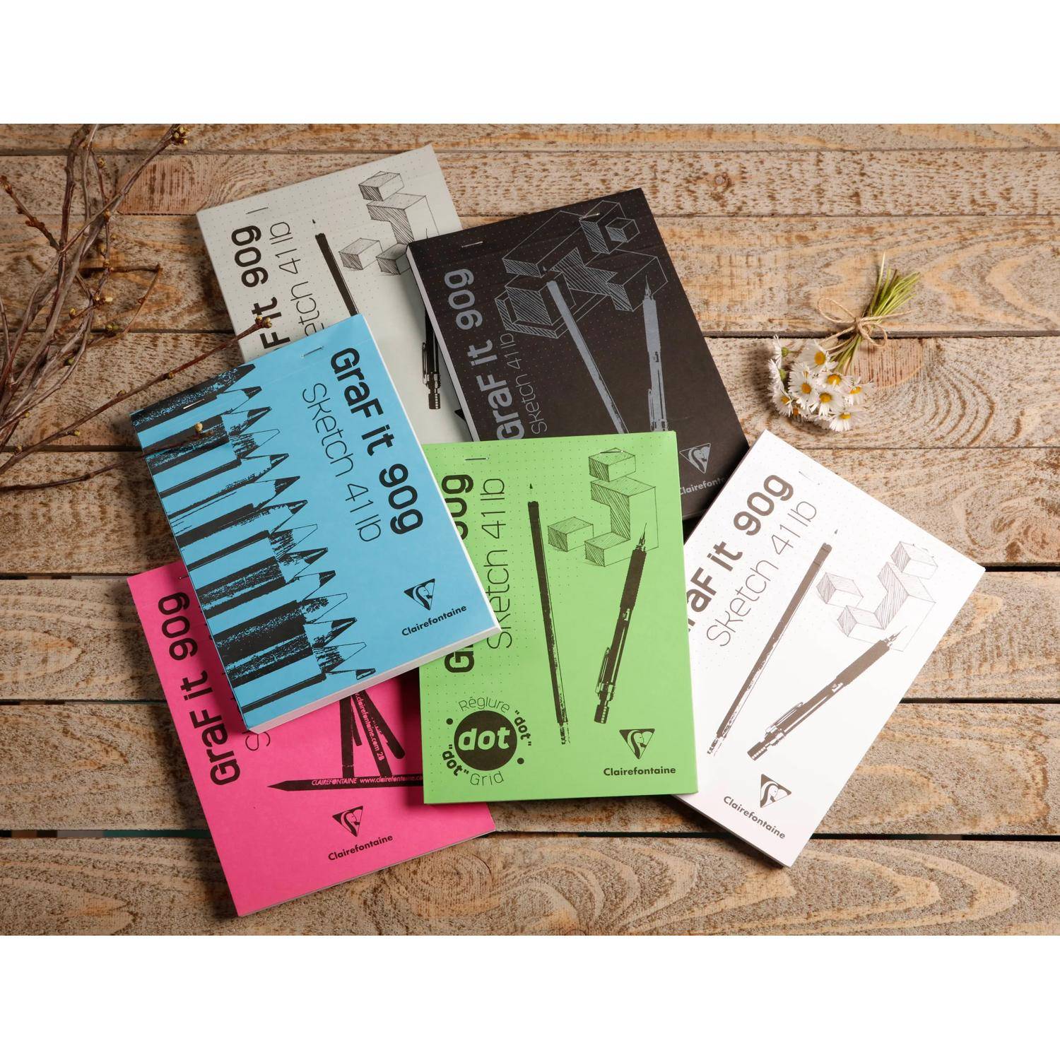 Assortierte "Graf It 909""-Skizzenbuch- und Zeichenblock-Umschläge in fünf Farben auf einem Holztisch arrangiert, mit einer kleinen Blume in der Nähe.