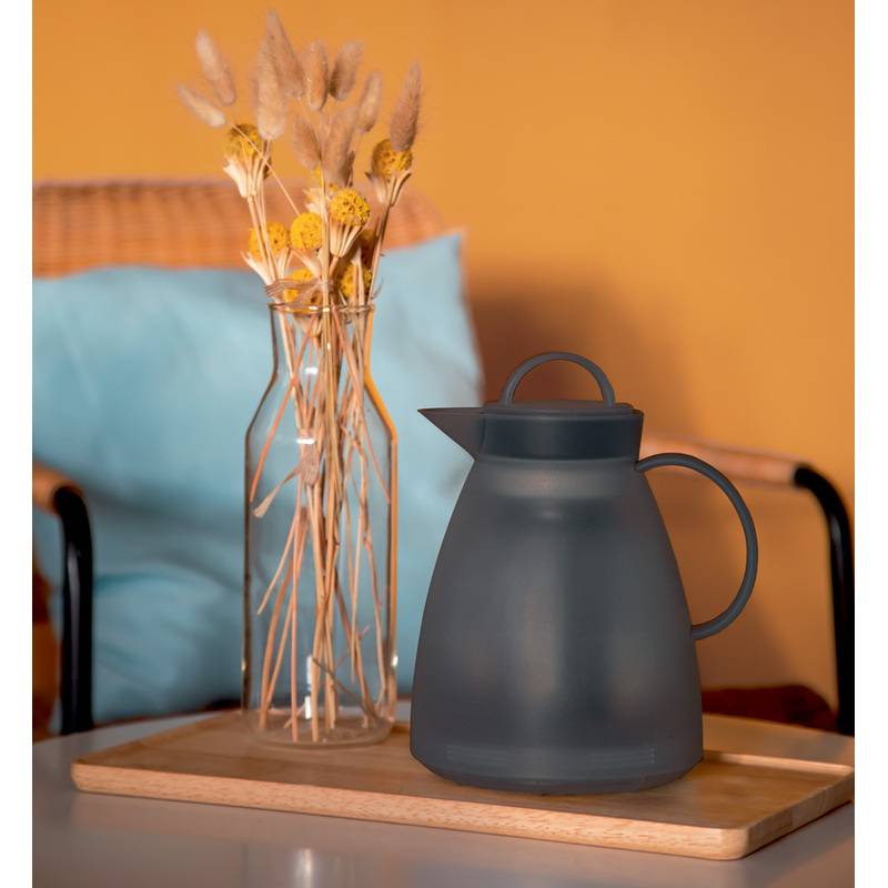 Ein matter schwarzer Wasserkocher steht auf einem Holztablett neben einer hohen Vase mit getrockneten gelben und beigen Blumen, vor einem weichen orange-blauen Hintergrund.