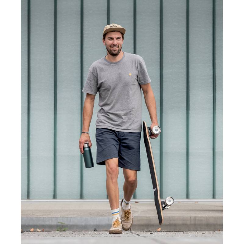 Ein Mann in lässiger Kleidung geht mit einem Skateboard in der einen und einer Wasserflasche in der anderen Hand vor einer urbanen Kulisse.