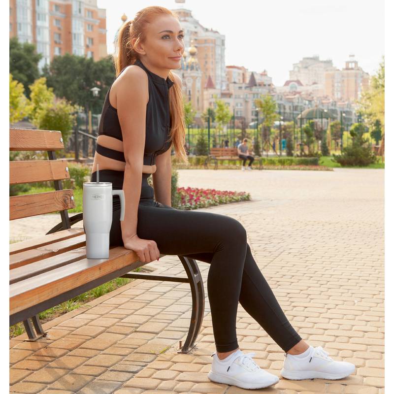 Eine Person sitzt auf einer Bank in einem Park, trägt Sportkleidung und hält einen Mixer mit einem Smoothie. Im Hintergrund sind Gebäude und Grünflächen zu sehen.