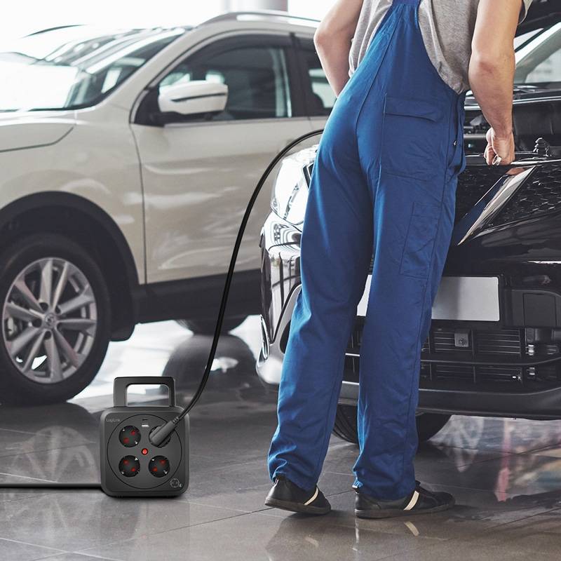 Ein Mechaniker in blauer Arbeitskleidung lädt ein Elektroauto mit einem Ladekabel. Daneben ist im Hintergrund ein weiteres Auto geparkt.