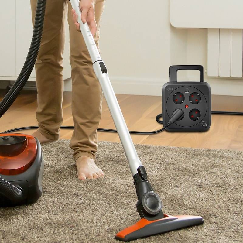 Eine Person, die einen Teppichboden mit einem Standstaubsauger in einem aufgeräumten Raum saugt. Ein Heizkörper und eine Steckdosenleiste sind im Hintergrund sichtbar.