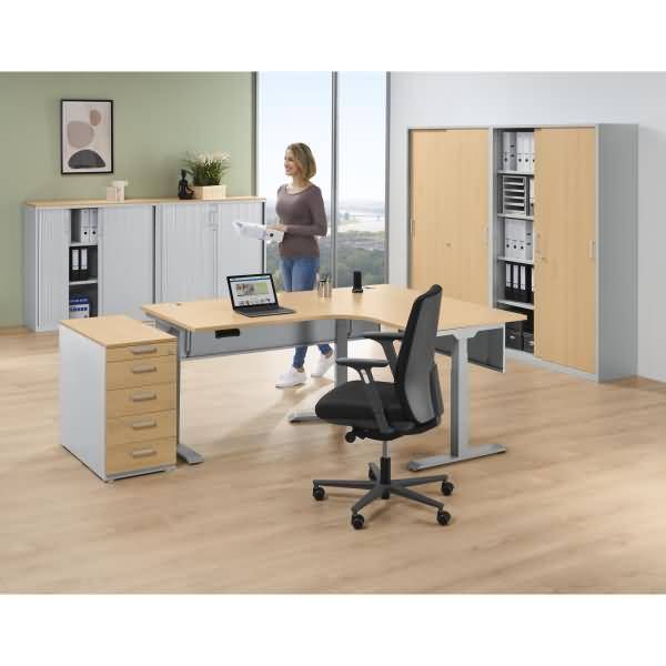 Modernes Büro mit Holzschreibtischen, Rollstuhl, Schränken, Laptop und einer Frau, die Papiere in der Nähe eines Fensters hält.