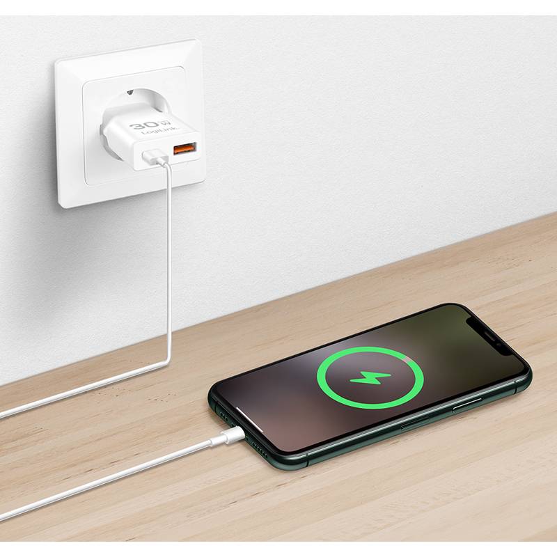 Ein Handy liegt auf einer Holzoberfläche und ist durch ein weißes Ladekabel mit einer Wandsteckdose verbunden. Der Bildschirm des Handys zeigt ein Ladesymbol.