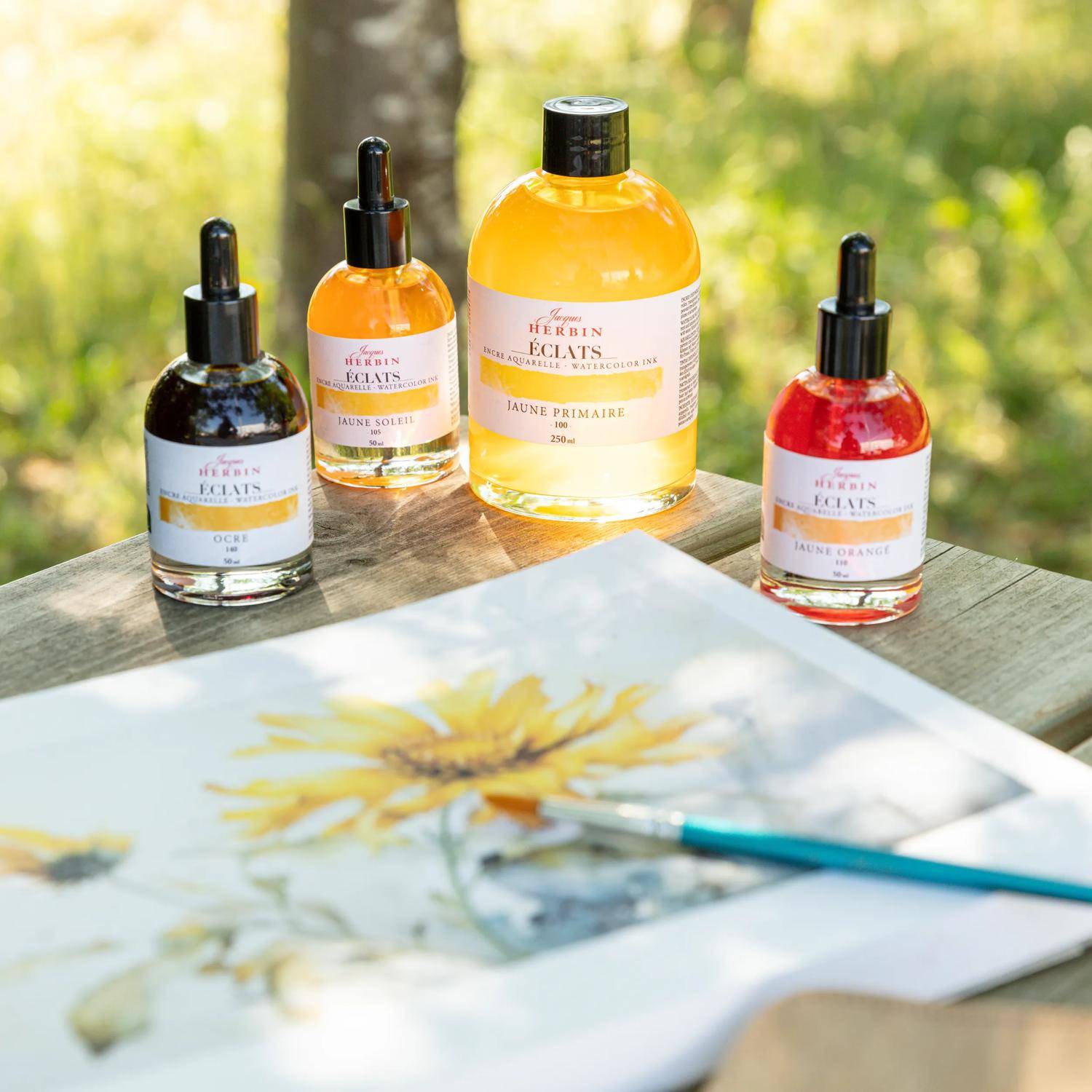 Tintenfläschchen in verschiedenen Farben stehen auf einem Holztisch im Freien neben einem Blumengemälde, während die Sonne durch die Bäume scheint.