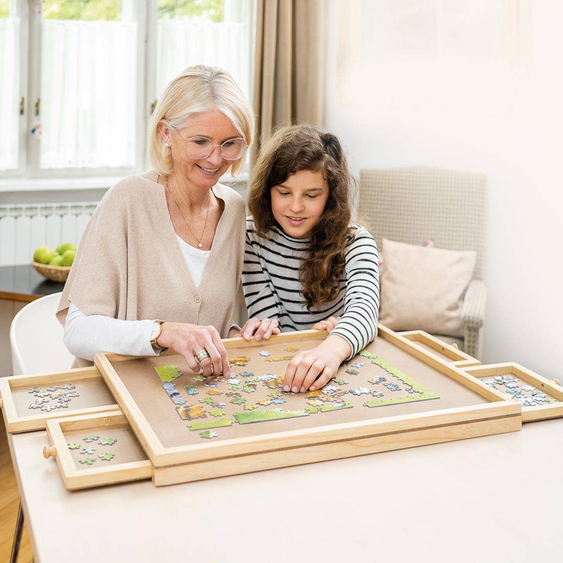 Eine lächelnde Frau und ein kleines Mädchen arbeiten zusammen an einem Puzzle auf einem Tisch in einem gemütlichen Zimmer und zeigen einen warmen, gemeinsamen Moment.