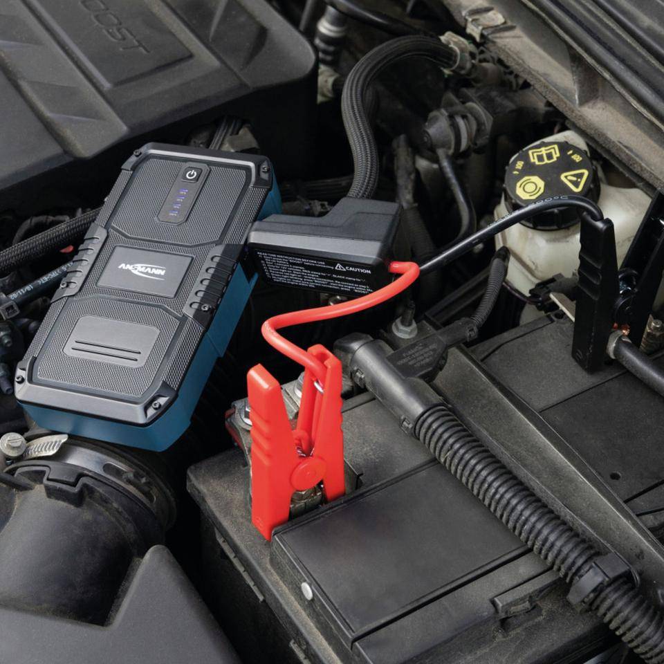 Tragbarer Starthilfegerät, das mit einer Autobatterie in einem Motorraum verbunden ist, mit roten und schwarzen Klemmen zum Starthilfen geben.