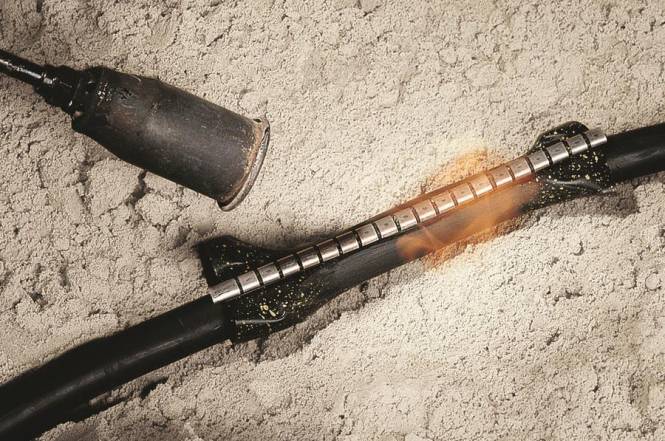 Ein glimmender Zigarillo neben einem gezahnten Metallrohr auf einer sandigen Oberfläche liegend, was auf kürzliche Verwendung oder weggeworfene Gegenstände hindeutet.