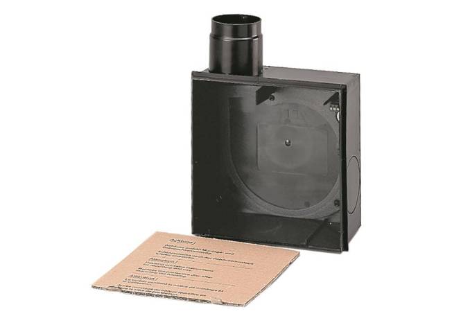 Eine schwarze Metallbox mit einer kreisförmigen Öffnung auf der Oberseite sitzt neben einem Kartonblatt, das Anweisungen oder Warnhinweise anzeigt, wahrscheinlich zur Installation.
