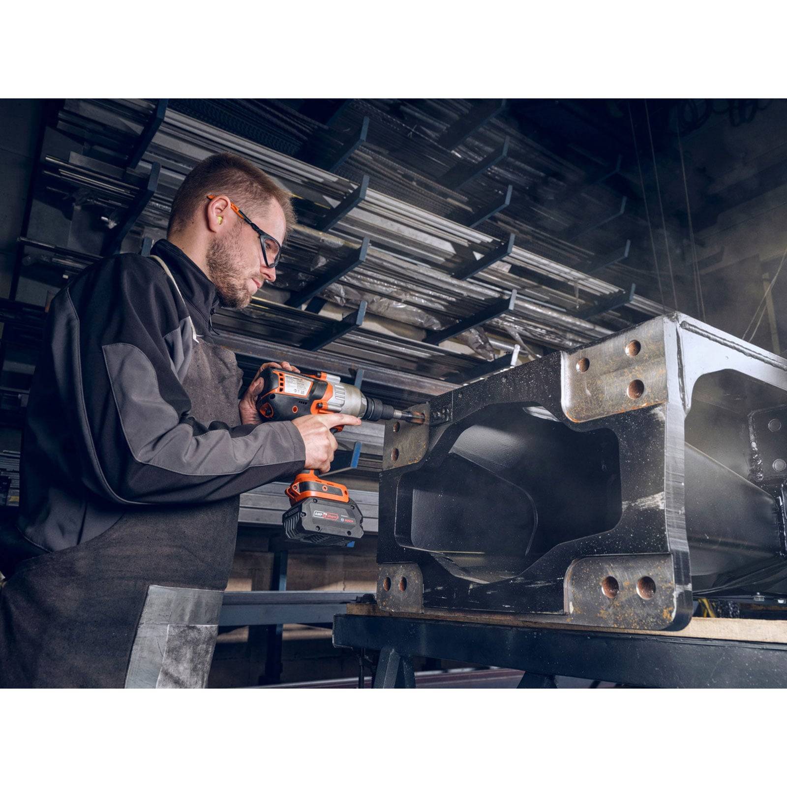 Ein Arbeiter in Schutzausrüstung verwendet eine Bohrmaschine an einem großen Metallrahmen in einem industriellen Werkstattumfeld.