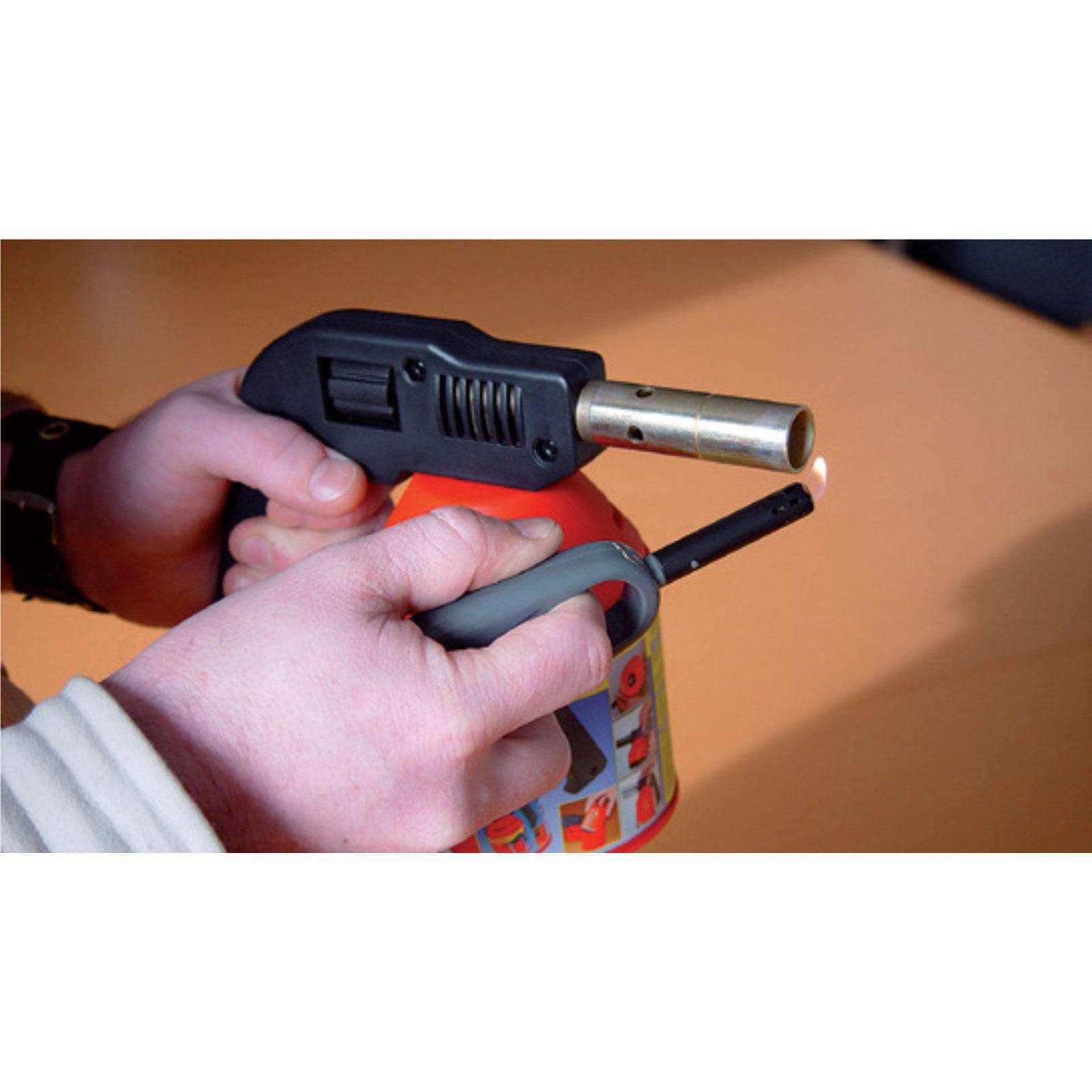 Eine Person zündet einen Gasbrenner an und hält ihn mit beiden Händen. Die Flamme ist klein, und der Brenner hat einen roten Behälter und eine schwarze Düse.