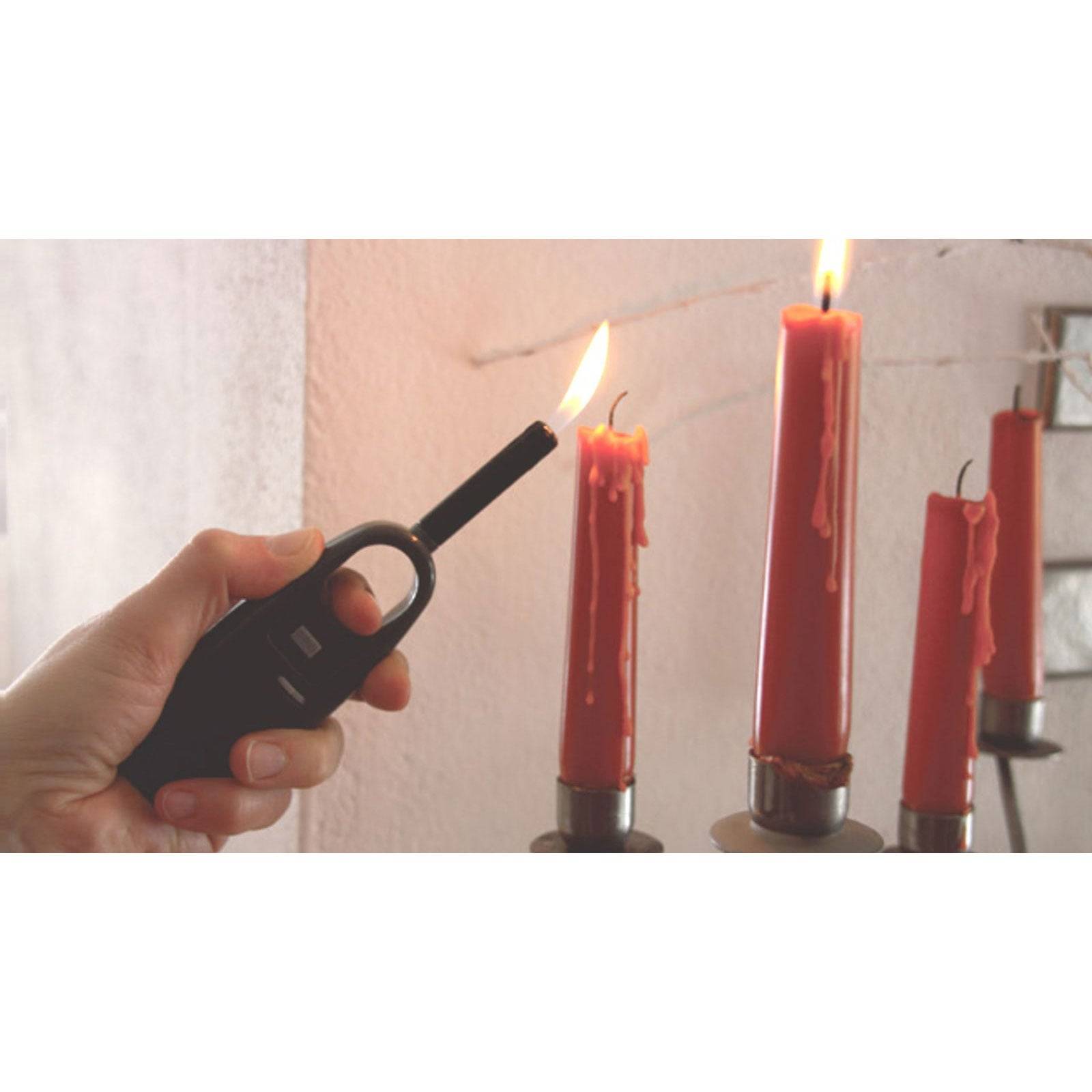 Eine Hand hält ein Feuerzeug und zündet eine rote Kerze auf einem Metallständer an, während zwei weitere rote Kerzen daneben bereits brennen.